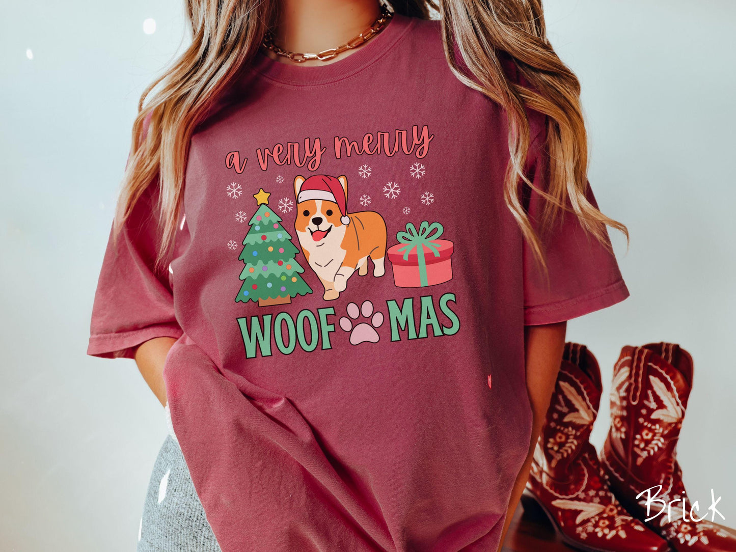 A vintage brick colored Comfort Colors T-shirt with the text 'a very merry Woofmas' in red and green font, featuring an orange corgi wearing a red Santa hat sitting between a Christmas tree and a present with snowflakes hanging in the air.