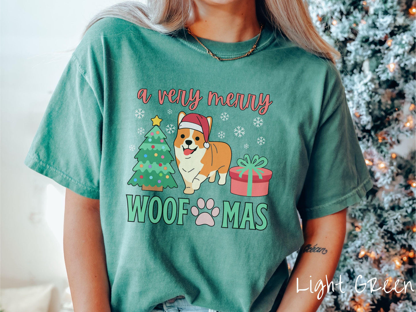 A vintage light green colored Comfort Colors T-shirt with the text 'a very merry Woofmas' in red and green font, featuring an orange corgi wearing a red Santa hat sitting between a Christmas tree and a present with snowflakes hanging in the air.