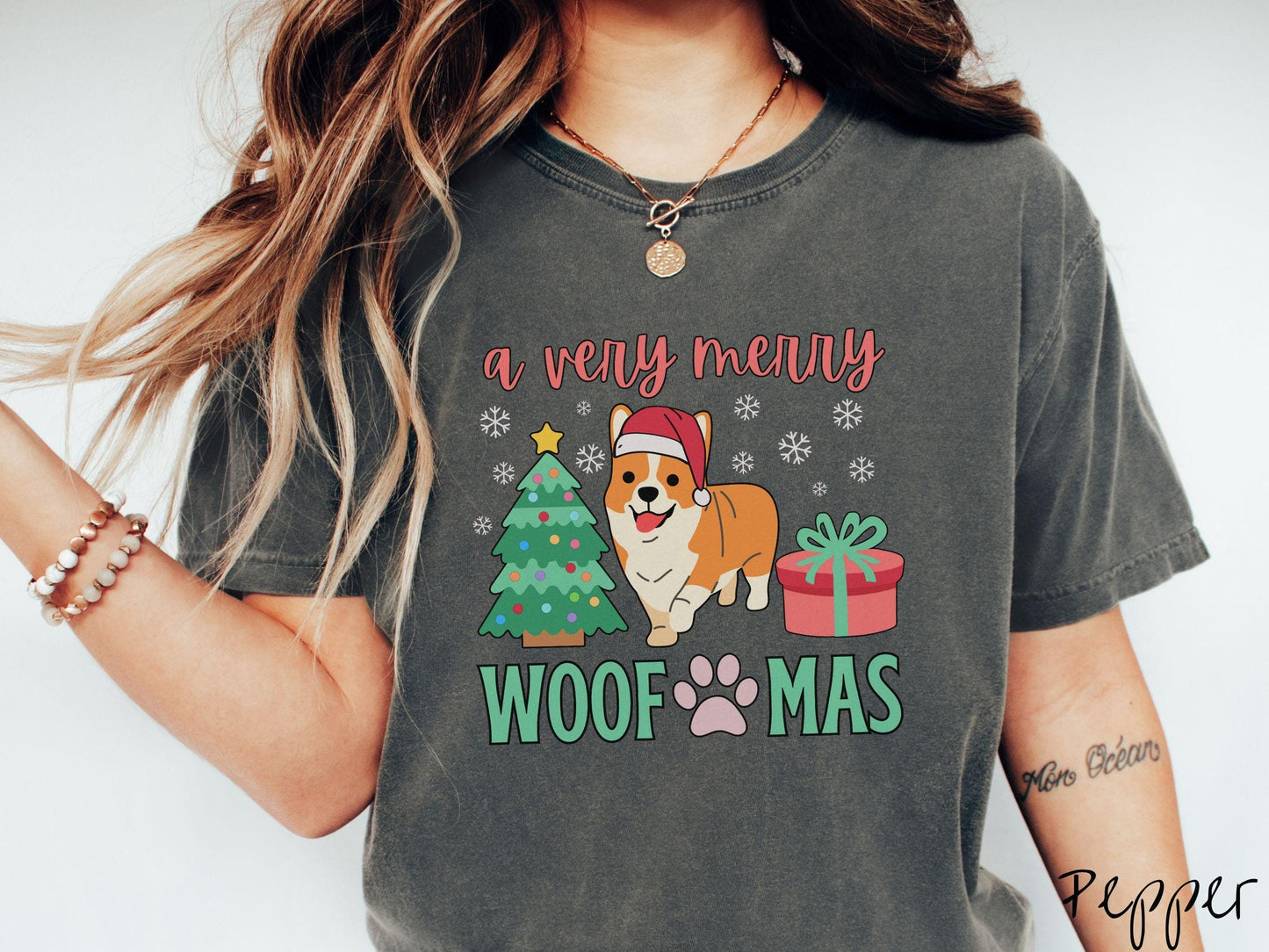 A vintage pepper colored Comfort Colors T-shirt with the text 'a very merry Woofmas' in red and green font, featuring an orange corgi wearing a red Santa hat sitting between a Christmas tree and a present with snowflakes hanging in the air.