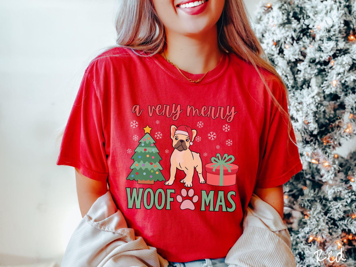A vintage red colored Comfort Colors T-shirt with the text 'a very merry Woofmas' in red and green font, featuring a French Bulldog wearing a red Santa hat sitting between a Christmas tree and a present with white snowflakes hanging in the air.