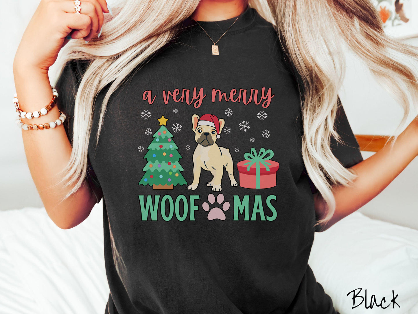 A vintage black colored Comfort Colors T-shirt with the text 'a very merry Woofmas' in red and green font, featuring a French Bulldog wearing a red Santa hat sitting between a Christmas tree and a present with white snowflakes hanging in the air.