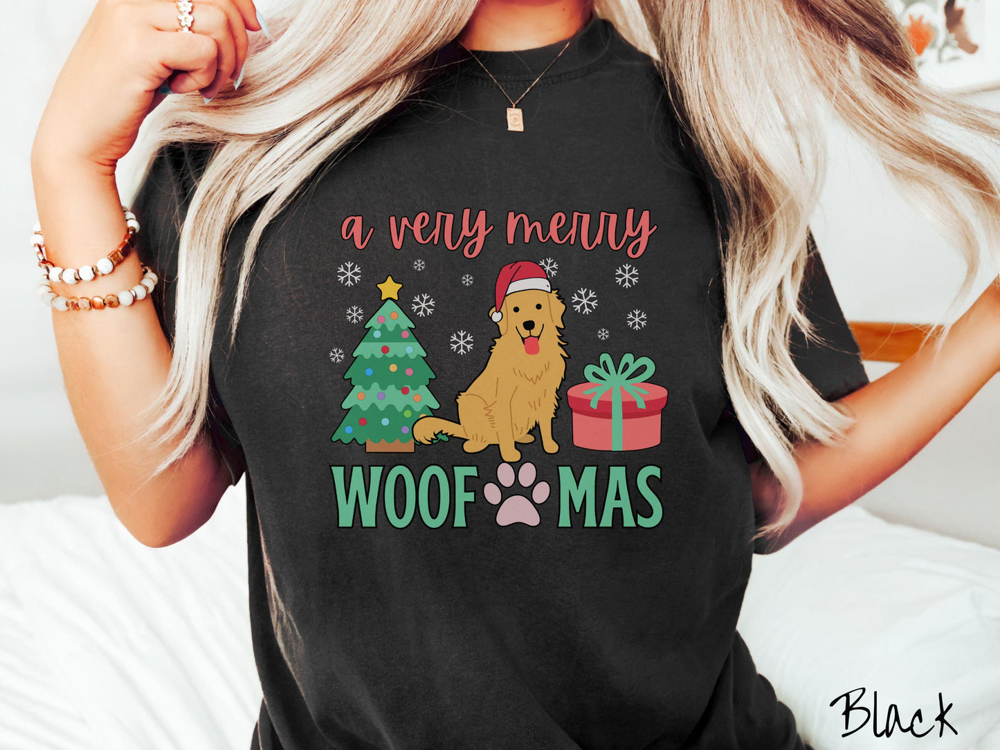 A vintage black colored Comfort Colors T-shirt with the text 'a very merry Woofmas' in red and green font, featuring a Golden Retriever wearing a red Santa hat sitting between a Christmas tree and a present with snowflakes hanging in the air.