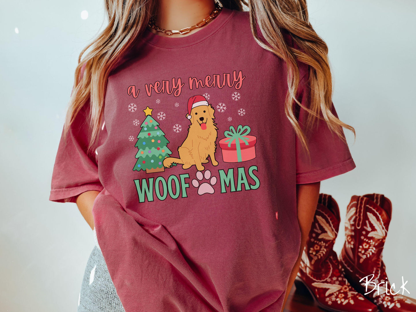 A vintage brick colored Comfort Colors T-shirt with the text 'a very merry Woofmas' in red and green font, featuring a Golden Retriever wearing a red Santa hat sitting between a Christmas tree and a present with snowflakes hanging in the air.