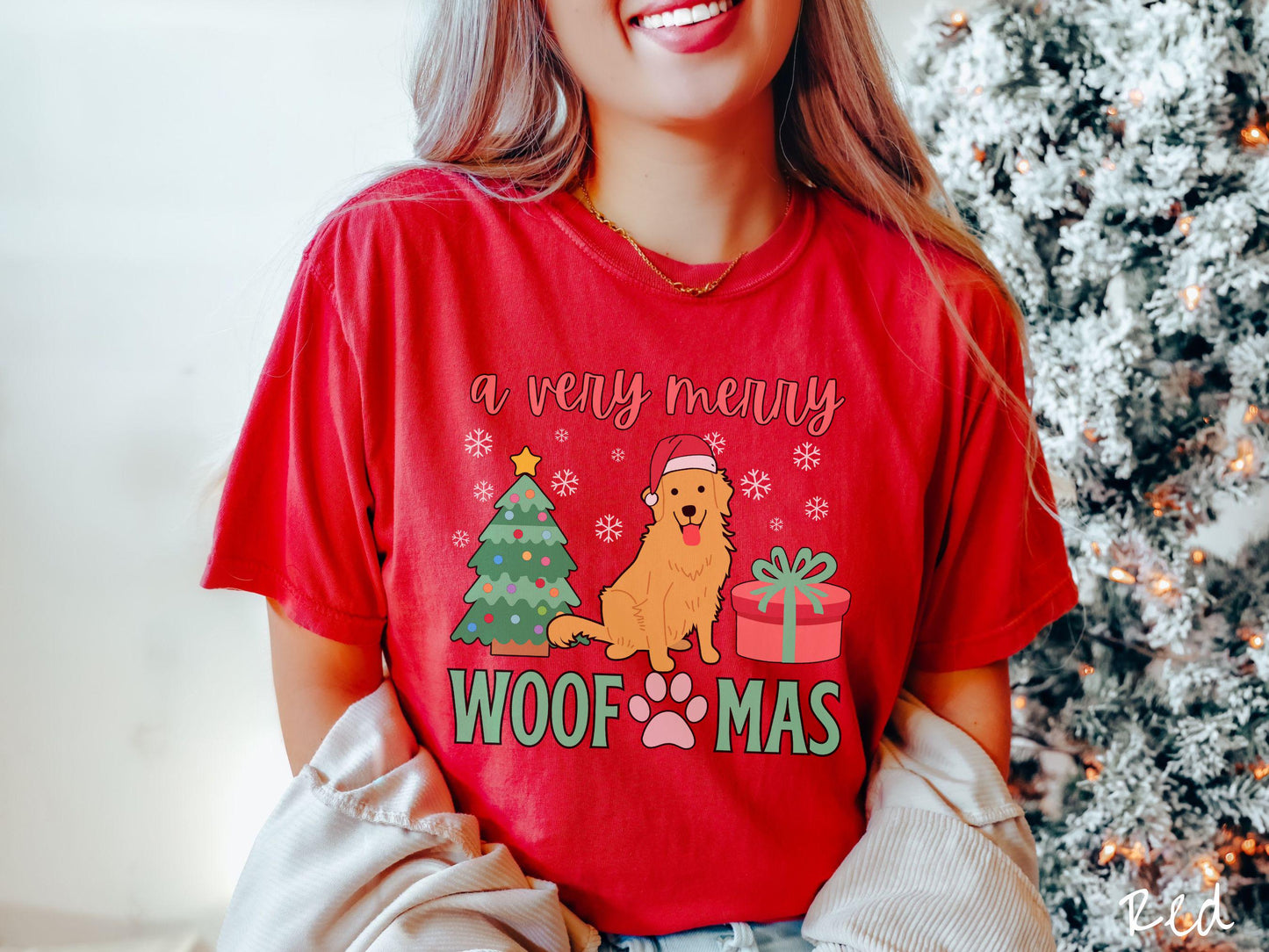 A vintage red colored Comfort Colors T-shirt with the text 'a very merry Woofmas' in red and green font, featuring a Golden Retriever wearing a red Santa hat sitting between a Christmas tree and a present with snowflakes hanging in the air.