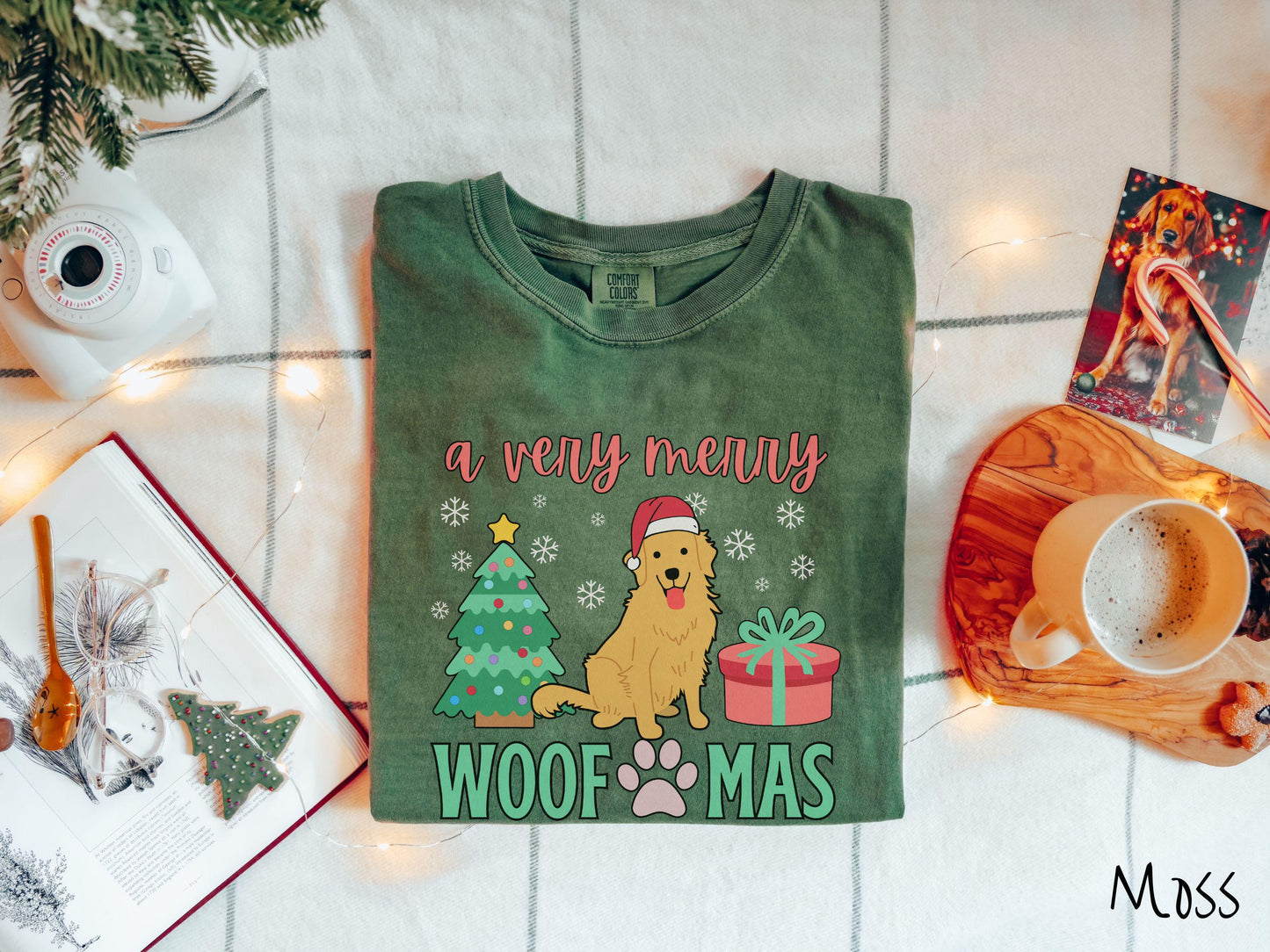 A vintage moss colored Comfort Colors T-shirt with the text 'a very merry Woofmas' in red and green font, featuring a Golden Retriever wearing a red Santa hat sitting between a Christmas tree and a present with snowflakes hanging in the air.