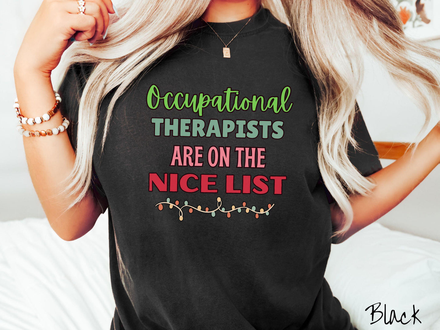 A vintage black colored Comfort Colors T-shirt features the text 'Occupational Therapy is on the nice list' in green, pink, and red fonts. There is a string of colorful Christmas lights along the bottom, exuding a cozy holiday vibe.
