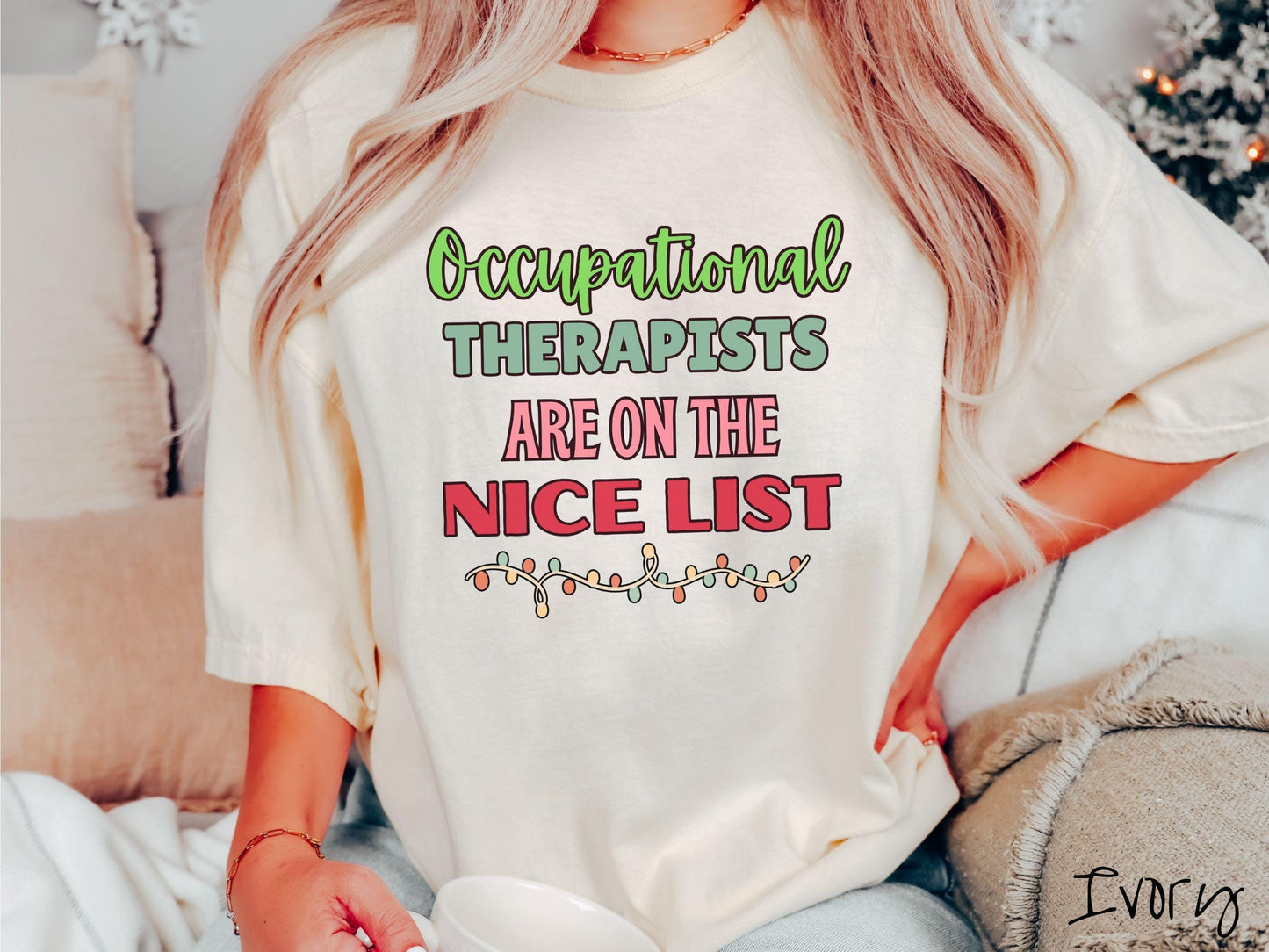 A vintage ivory colored Comfort Colors T-shirt features the text 'Occupational Therapy is on the nice list' in green, pink, and red fonts. There is a string of colorful Christmas lights along the bottom, exuding a cozy holiday vibe.