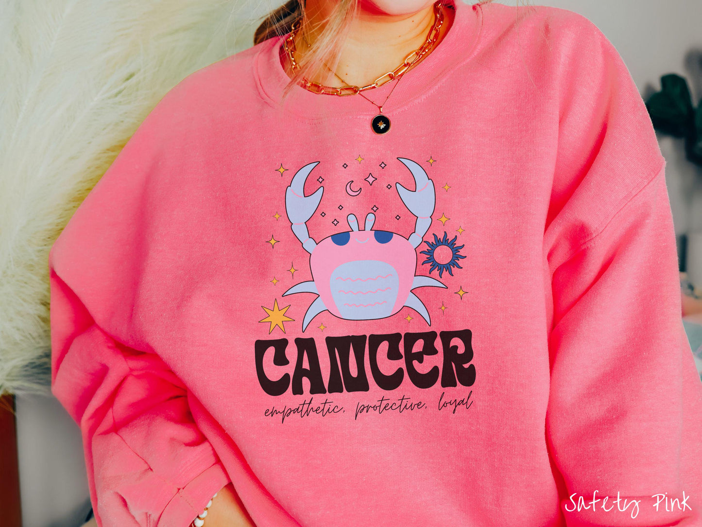 A vintage safety pink colored Comfort Colors sweatshirt features a large pink and blue crab, surrounded by stars, with the text Cancer and the words empathetic, protective, loyal.