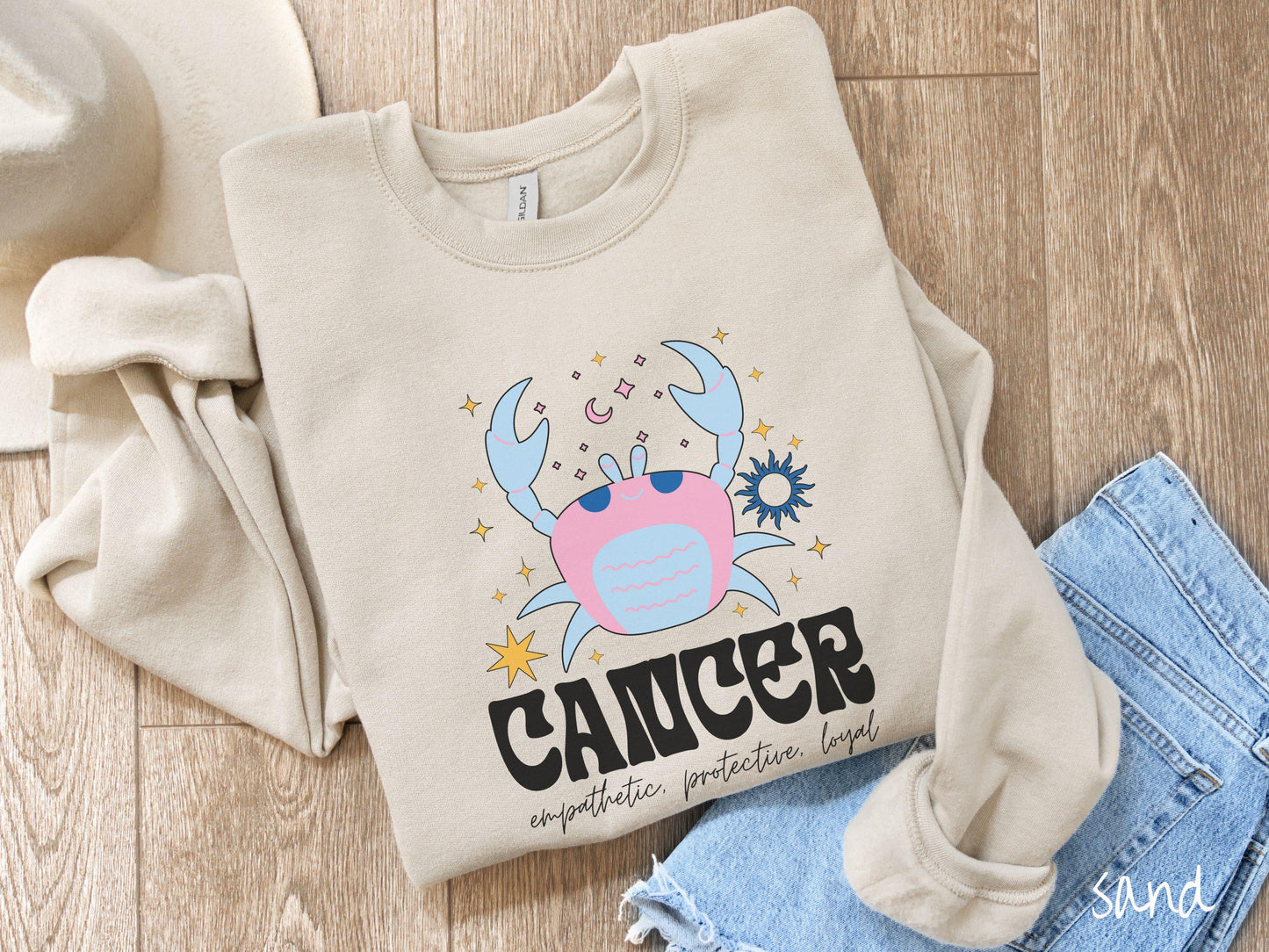 A vintage sand colored Comfort Colors sweatshirt features a large pink and blue crab, surrounded by stars, with the text Cancer and the words empathetic, protective, loyal.