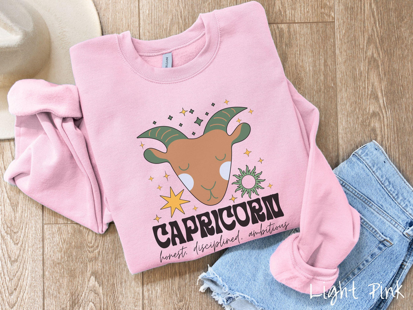 A vintage light pink colored Gildan sweatshirt features a large brown and green goat head surrounded by stars and the sun with the text Capricorn in large bold font and below that are the words honest, disciplined, ambitious in cursive font.