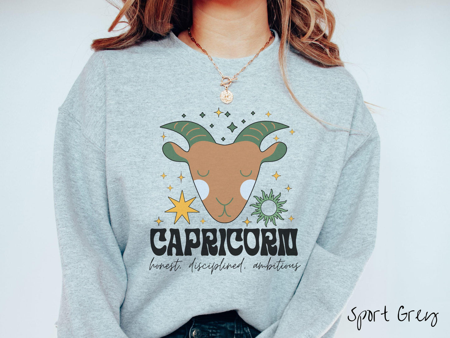 A vintage sport grey colored Gildan sweatshirt features a large brown and green goat head surrounded by stars and the sun with the text Capricorn in large bold font and below that are the words honest, disciplined, ambitious in cursive font.