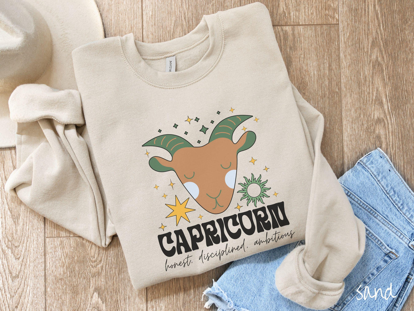 A vintage sand colored Gildan sweatshirt features a large brown and green goat head surrounded by stars and the sun with the text Capricorn in large bold font and below that are the words honest, disciplined, ambitious in cursive font.