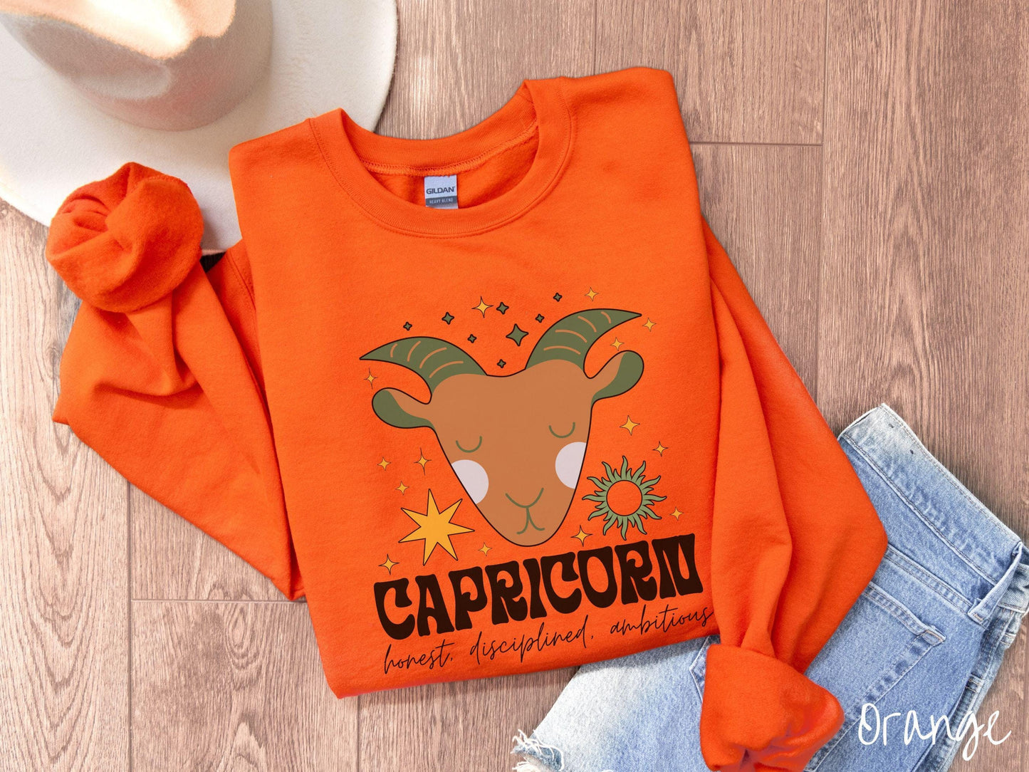 A vintage orange colored Gildan sweatshirt features a large brown and green goat head surrounded by stars and the sun with the text Capricorn in large bold font and below that are the words honest, disciplined, ambitious in cursive font.