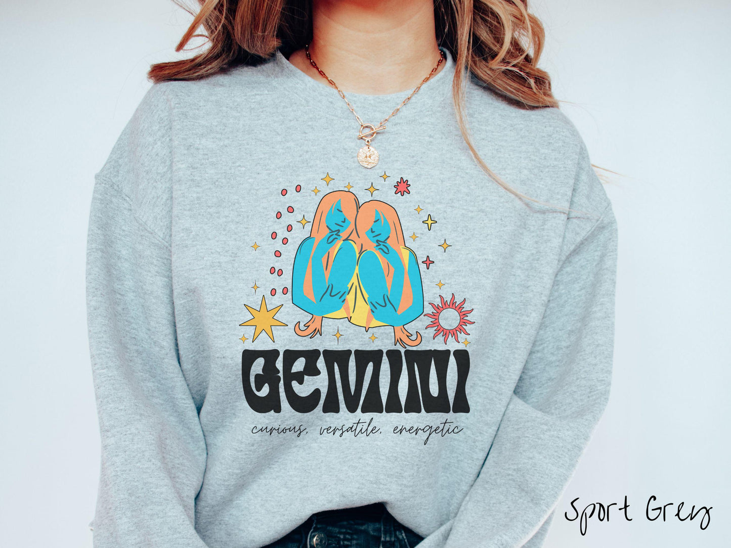 A vintage sport grey colored Gildan sweatshirt features the twins Castor and Pollux in blue and orange colors surrounded by stars and sun with text Gemini in large bold font and below that are the words curious, versatile, energetic in cursive font.