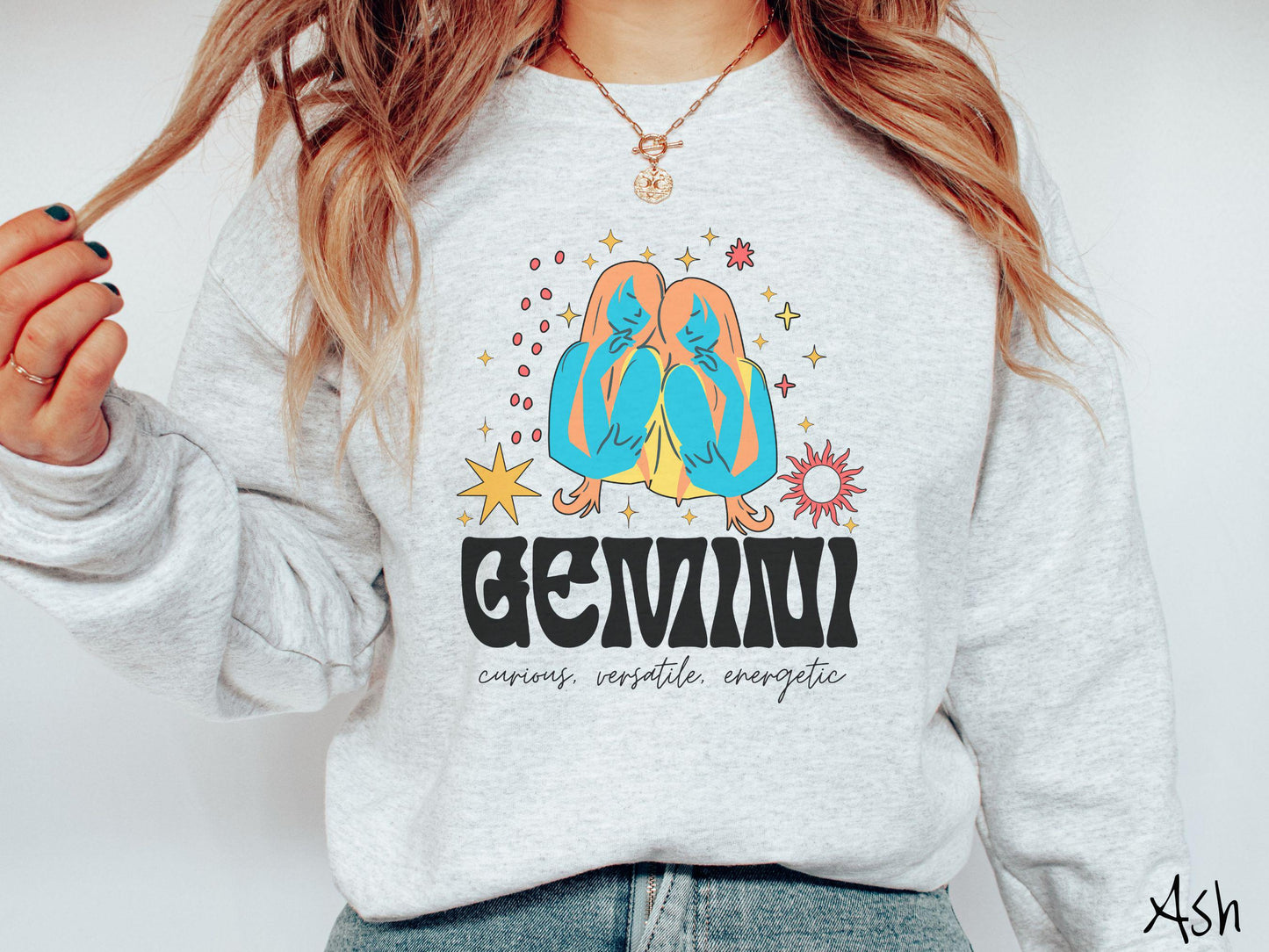A vintage ash colored Gildan sweatshirt features the twins Castor and Pollux in blue and orange colors surrounded by stars and sun with text Gemini in large bold font and below that are the words curious, versatile, energetic in cursive font.
