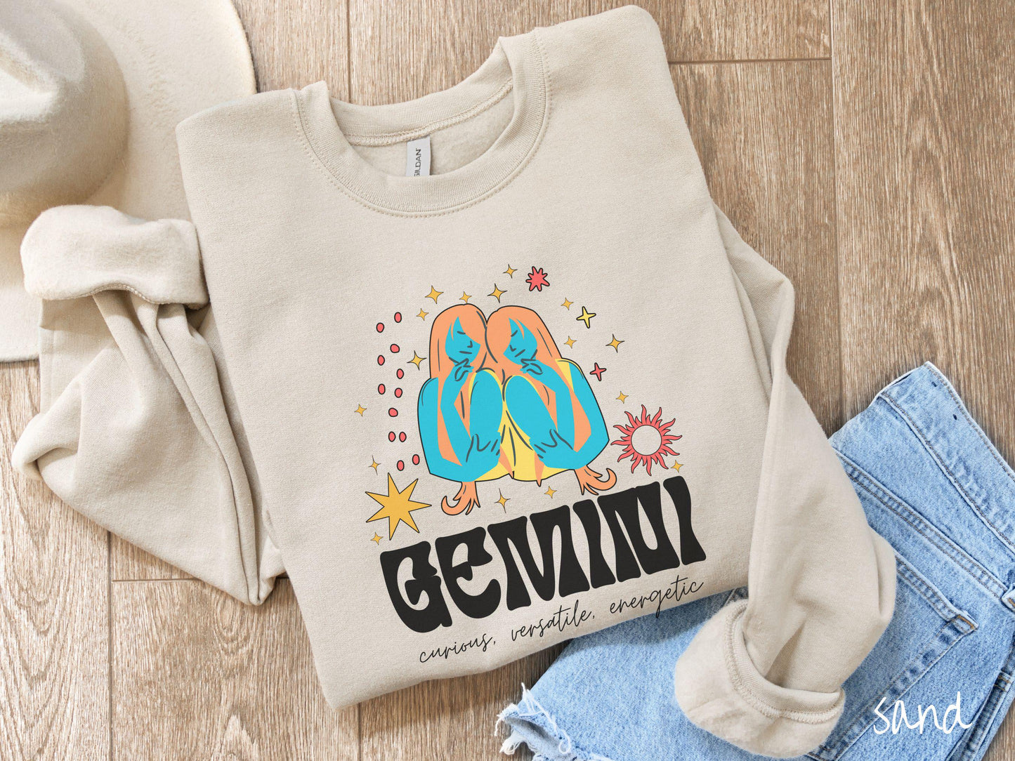 A vintage sand colored Gildan sweatshirt features the twins Castor and Pollux in blue and orange colors surrounded by stars and sun with text Gemini in large bold font and below that are the words curious, versatile, energetic in cursive font.