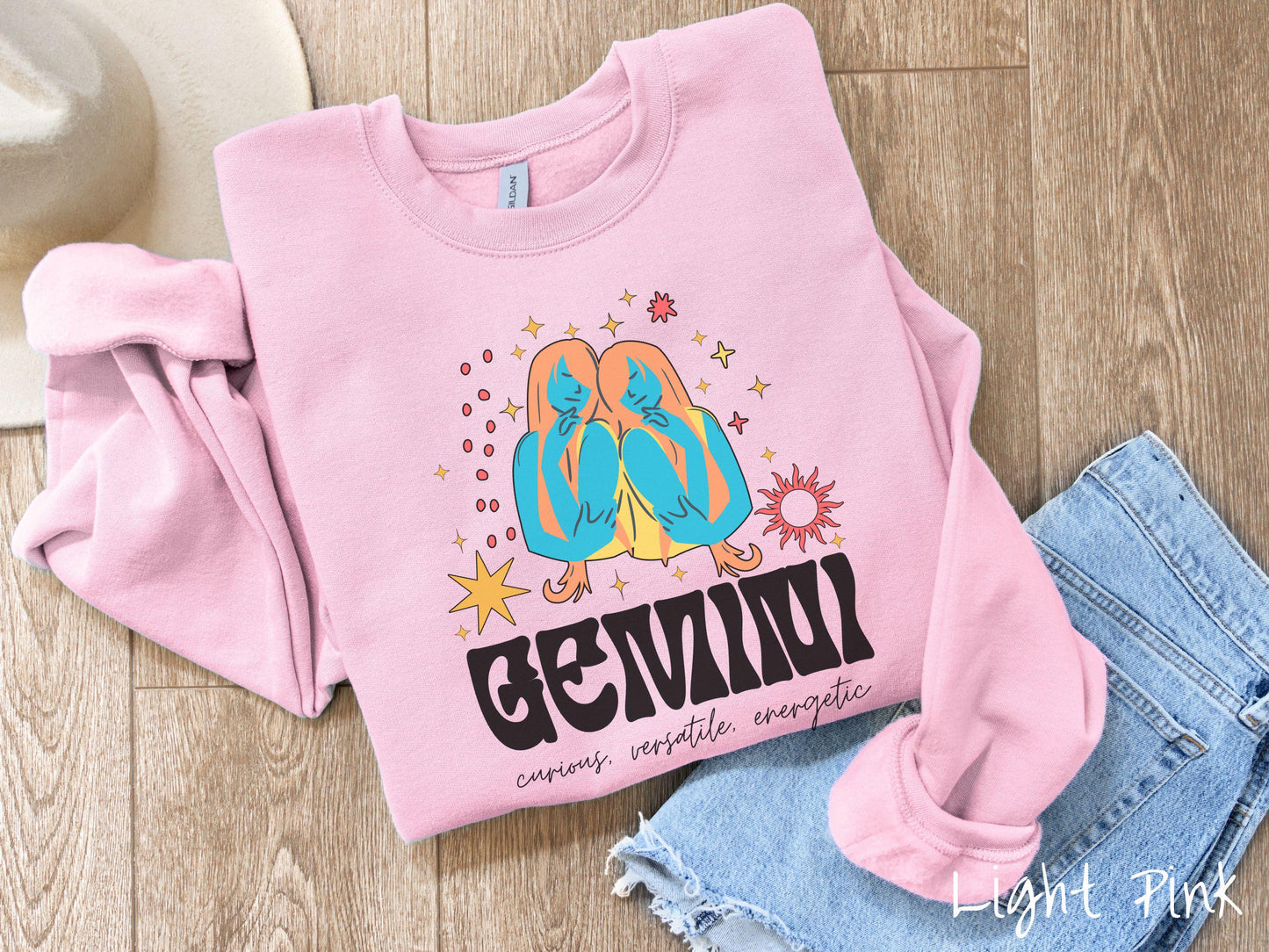 A vintage light pink colored Gildan sweatshirt features the twins Castor and Pollux in blue and orange colors surrounded by stars and sun with text Gemini in large bold font and below that are the words curious, versatile, energetic in cursive font.