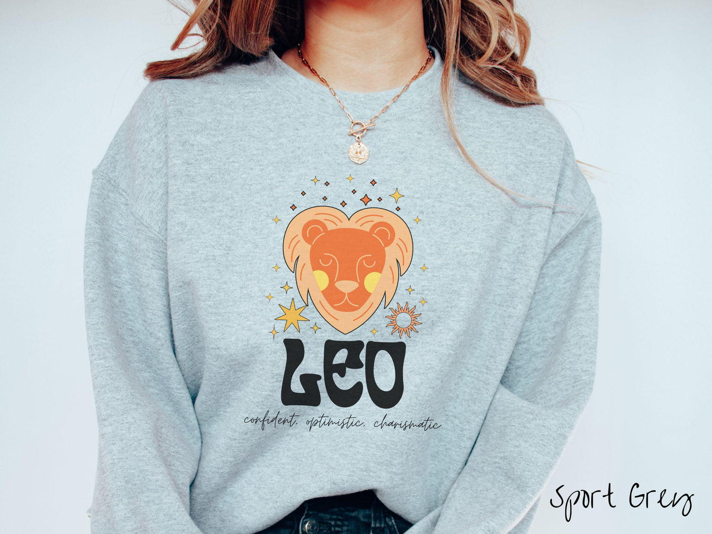 A vintage spot grey colored Gildan sweatshirt features a large orange and yellow lion surrounded by stars and the sun with the text Leo in large bold font and below that are the words confident, optimistic, charismatic in cursive font.