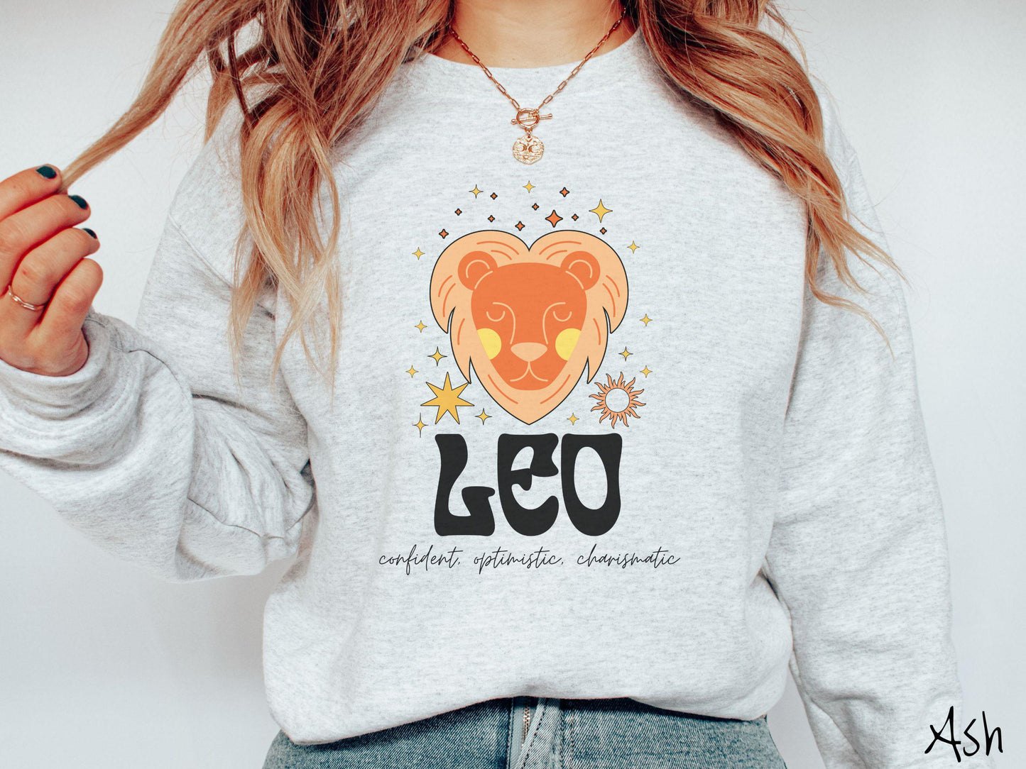 A vintage ash colored Gildan sweatshirt features a large orange and yellow lion surrounded by stars and the sun with the text Leo in large bold font and below that are the words confident, optimistic, charismatic in cursive font.