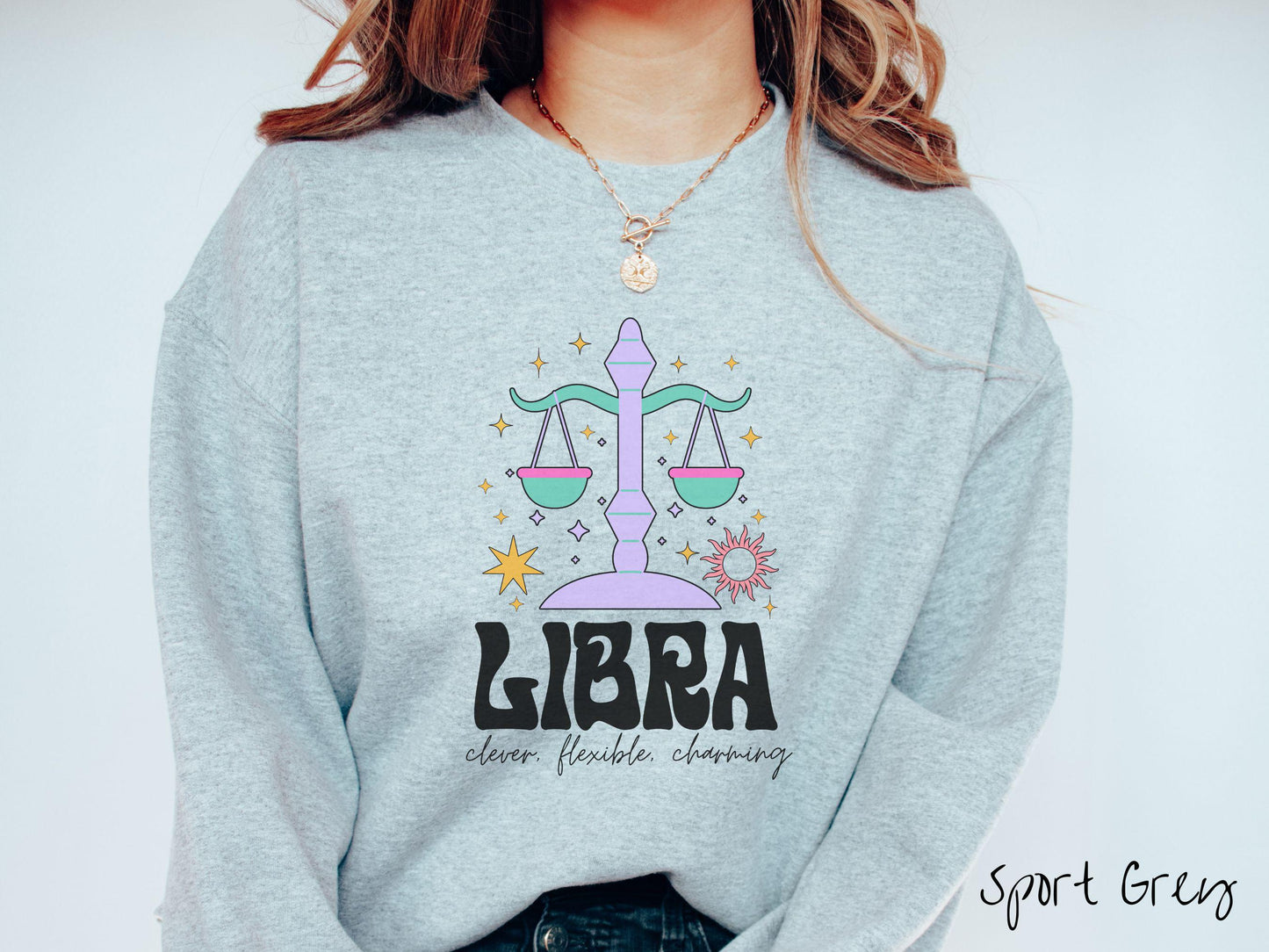 A vintage sport grey colored Gildan sweatshirt features a large purple and green weight scale surrounded by stars and the sun with the text Libra in large bold font and below that are the words clever, flexible, charming in cursive font.