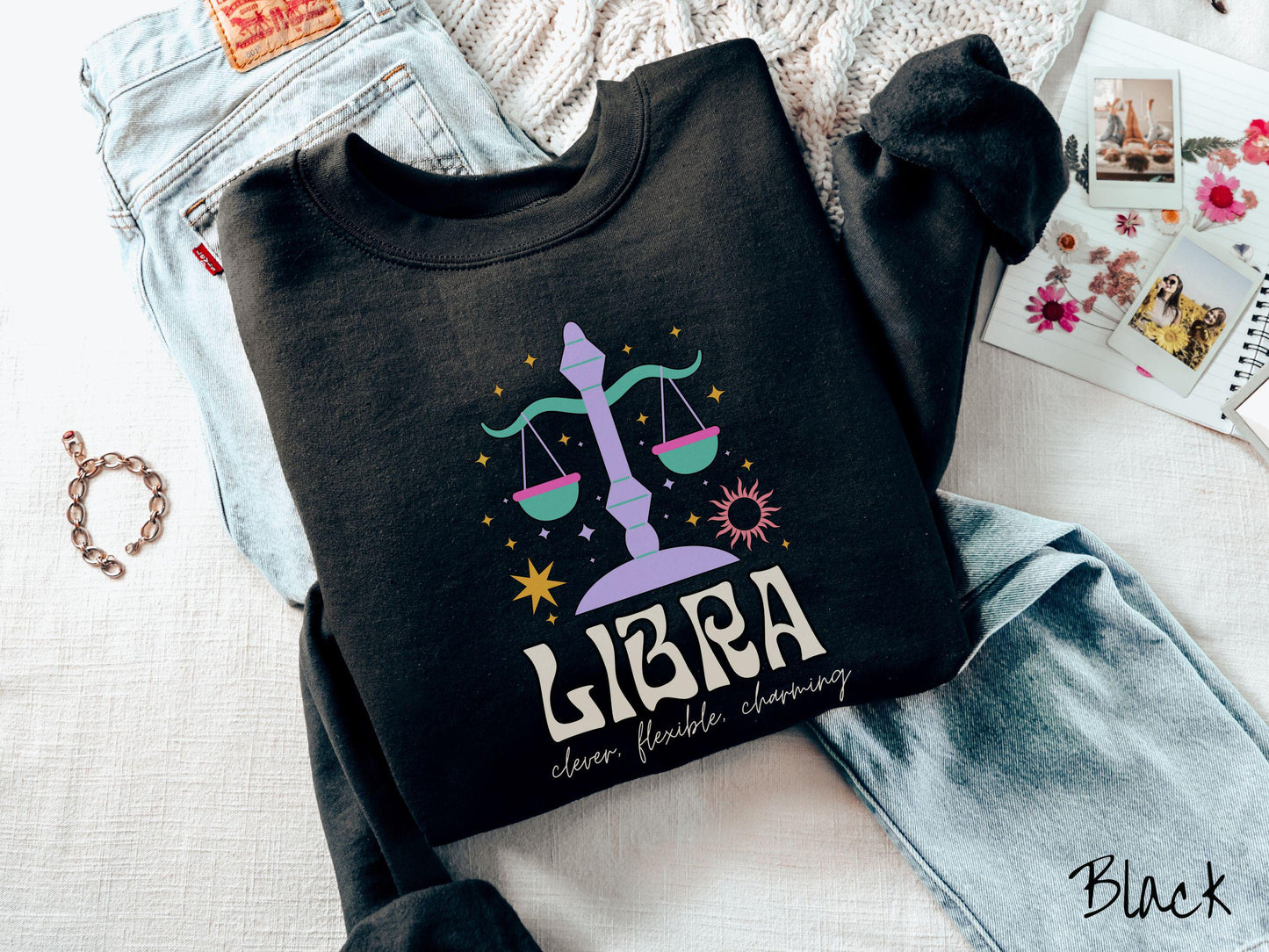 A vintage black colored Gildan sweatshirt features a large purple and green weight scale surrounded by stars and the sun with the text Libra in large bold font and below that are the words clever, flexible, charming in cursive font.