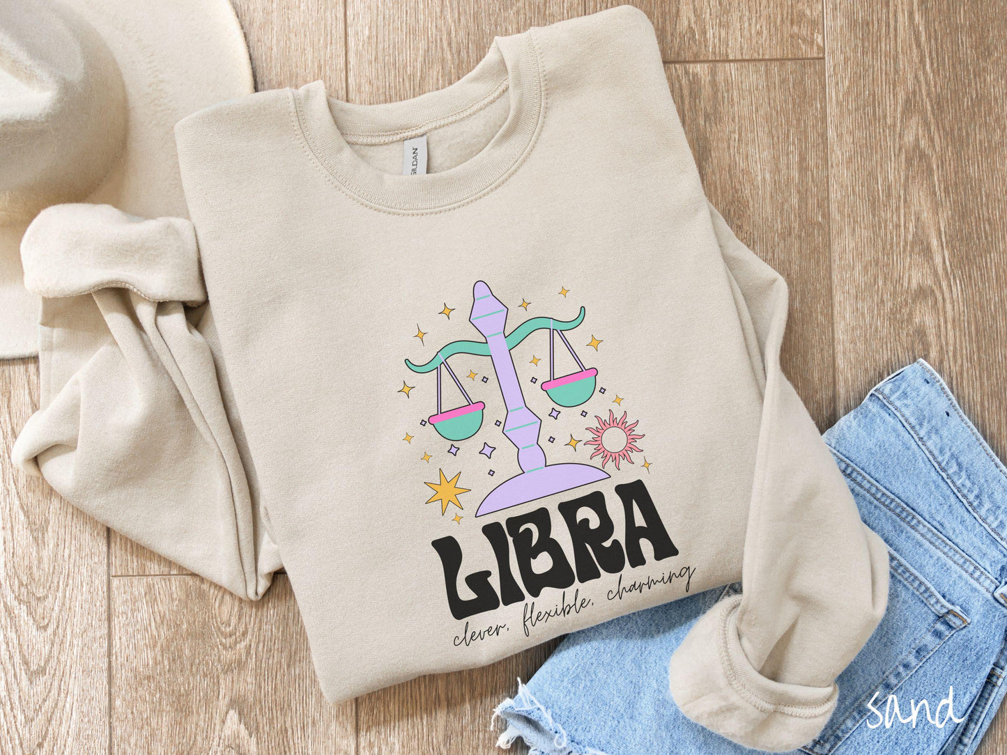 A vintage sand colored Gildan sweatshirt features a large purple and green weight scale surrounded by stars and the sun with the text Libra in large bold font and below that are the words clever, flexible, charming in cursive font.