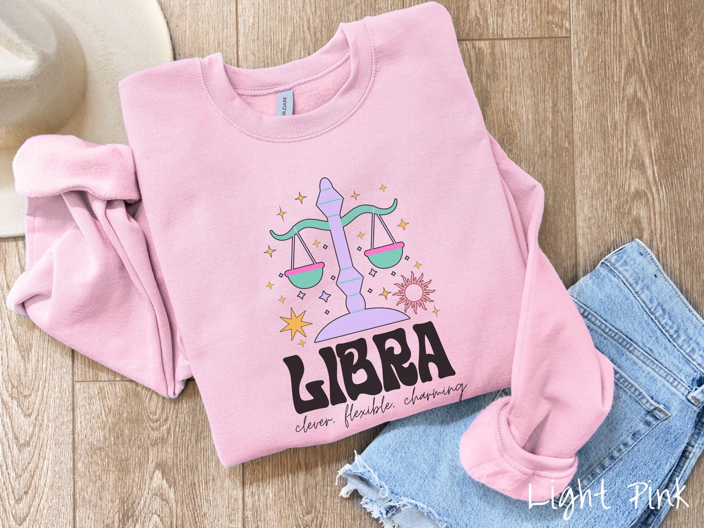 A vintage light pink colored Gildan sweatshirt features a large purple and green weight scale surrounded by stars and the sun with the text Libra in large bold font and below that are the words clever, flexible, charming in cursive font.