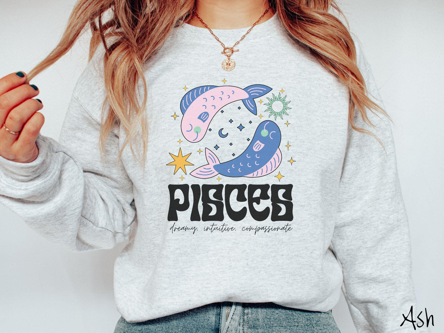A vintage ash colored Gildan sweatshirt features two large pink and blue fish surrounded by stars and the sun with the text Pisces in large bold font and below that are the words dreamy, intuitive, compassionate in cursive font.