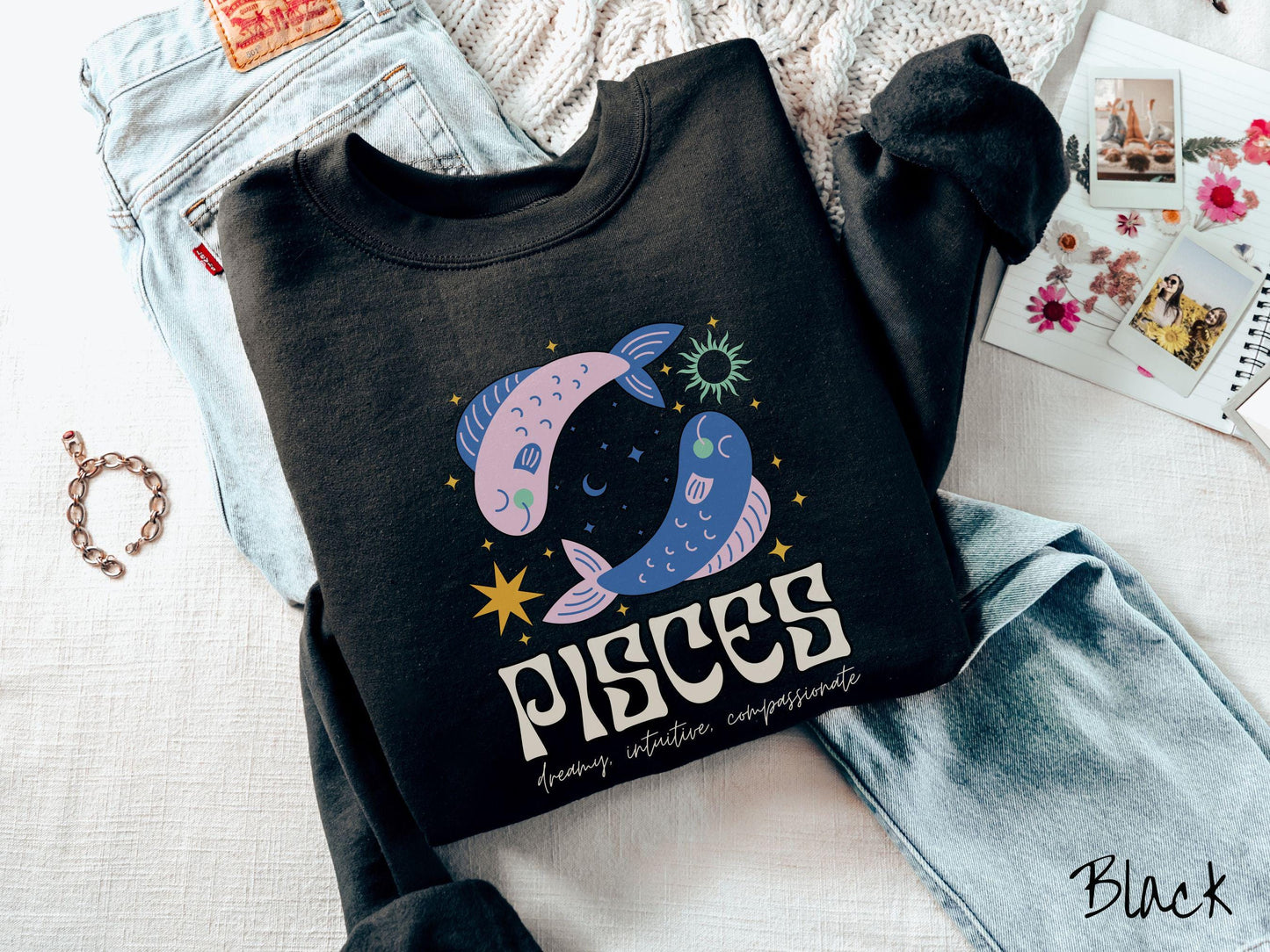 A vintage black colored Gildan sweatshirt features two large pink and blue fish surrounded by stars and the sun with the text Pisces in large bold font and below that are the words dreamy, intuitive, compassionate in cursive font.