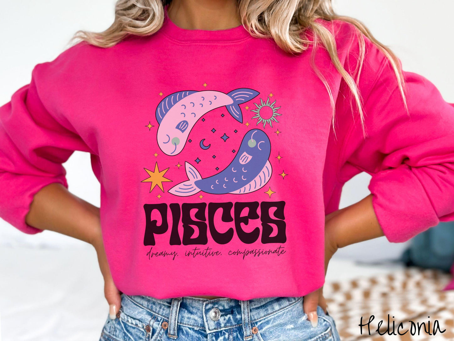 A vintage heliconia colored Gildan sweatshirt features two large pink and blue fish surrounded by stars and the sun with the text Pisces in large bold font and below that are the words dreamy, intuitive, compassionate in cursive font.