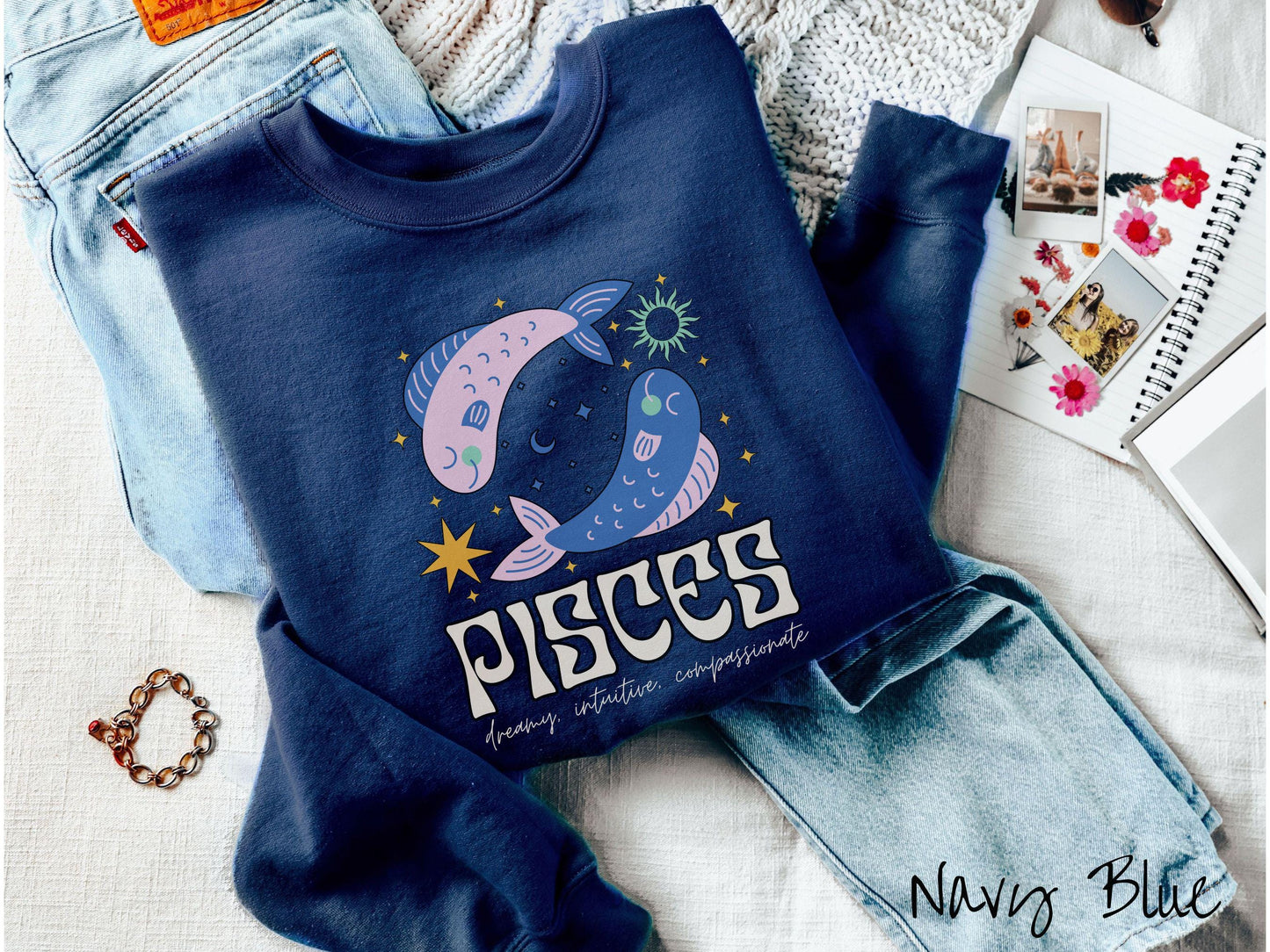 A vintage navy blue colored Gildan sweatshirt features two large pink and blue fish surrounded by stars and the sun with the text Pisces in large bold font and below that are the words dreamy, intuitive, compassionate in cursive font.