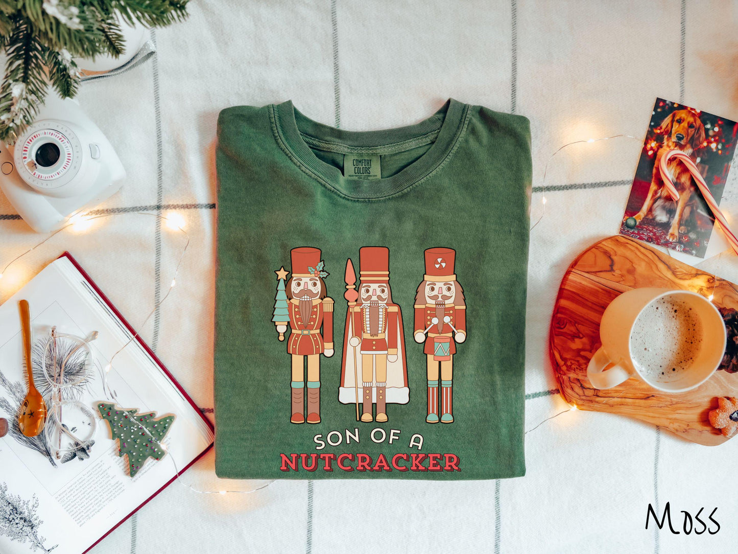 This vintage moss colored Comfort Colors T-shirt features three red nutcracker figurines dressed in Christmas attire and one cracking a nut. Below is the text 'Son of a nutcracker' in red and white font.