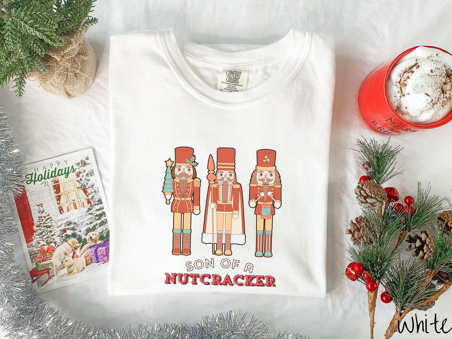 This vintage white colored Comfort Colors T-shirt features three red nutcracker figurines dressed in Christmas attire and one cracking a nut. Below is the text 'Son of a nutcracker' in red and white font.