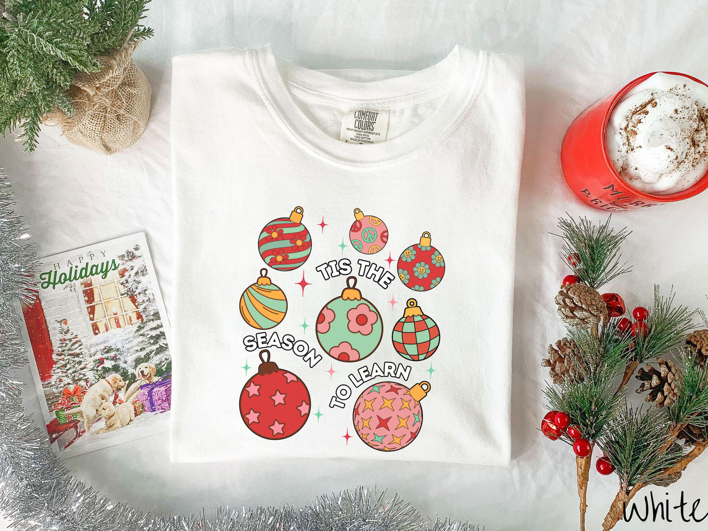 This vintage white colored Comfort Colors T-shirt features eight colorful Christmas tree ornaments arranged in a grid pattern and twinkling stars with the text Tis the Season to Learn.