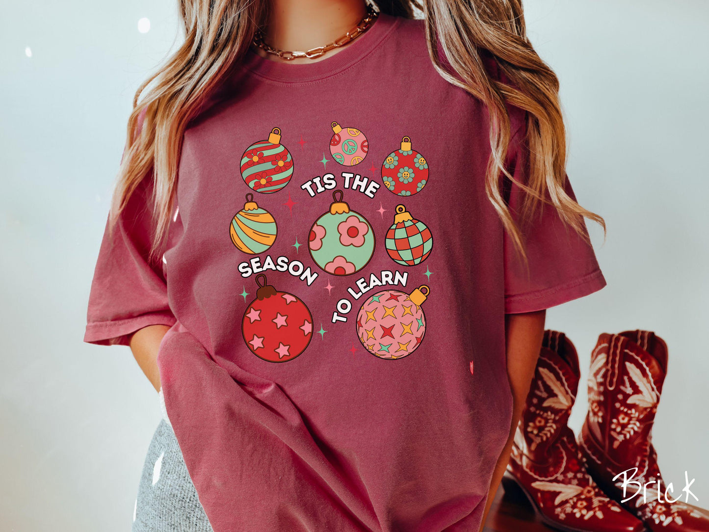 This vintage brick colored Comfort Colors T-shirt features eight colorful Christmas tree ornaments arranged in a grid pattern and twinkling stars with the text Tis the Season to Learn.