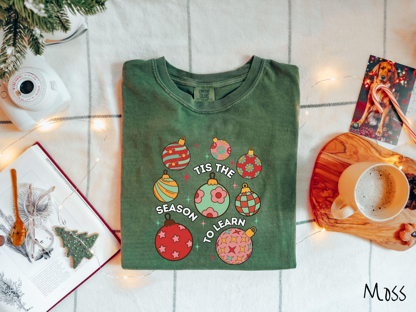 This vintage moss colored Comfort Colors T-shirt features eight colorful Christmas tree ornaments arranged in a grid pattern and twinkling stars with the text Tis the Season to Learn.