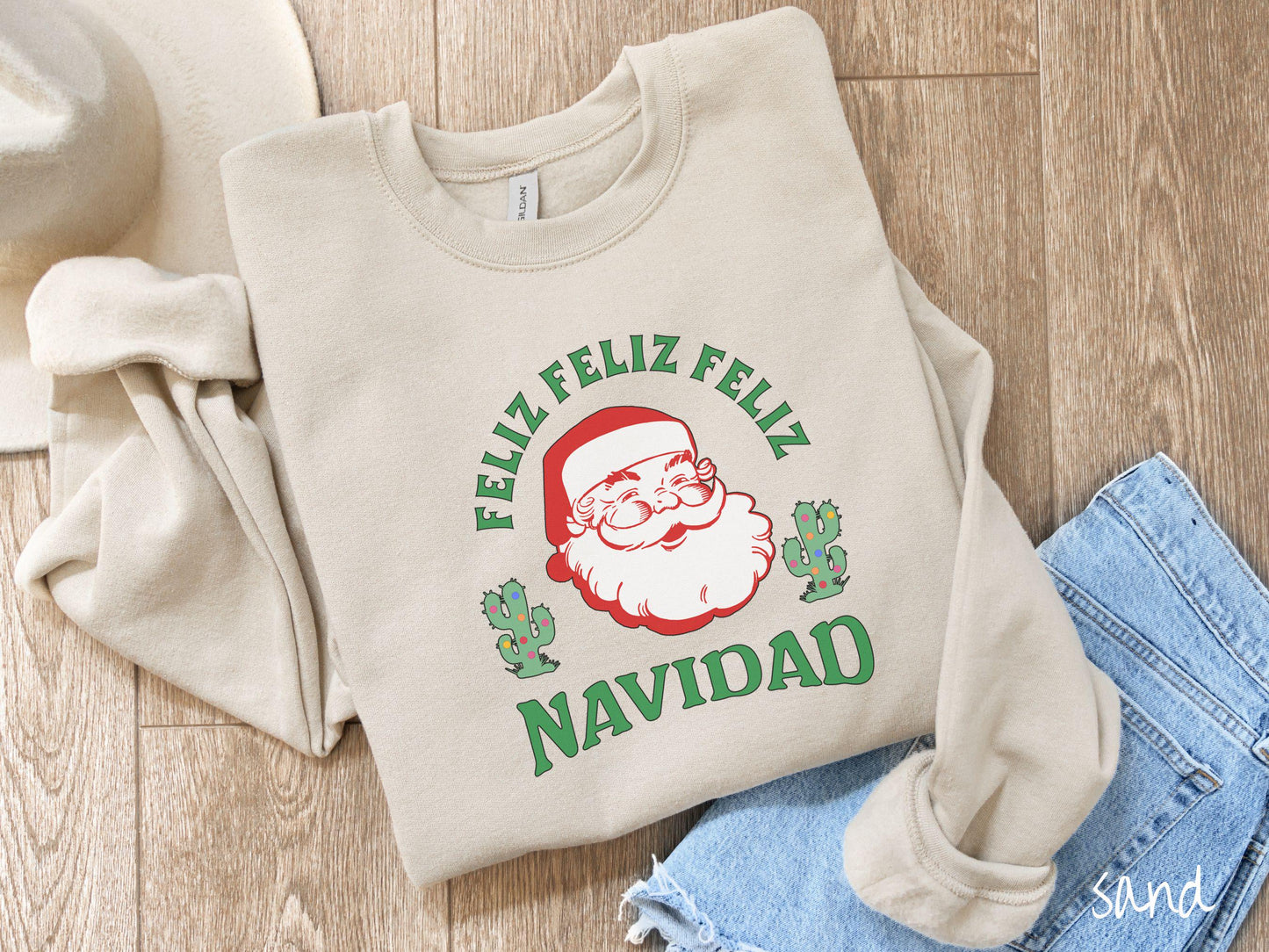 A festive sand colored Gildan sweatshirt featuring a smiling santa clause between two green cacti covered in colorful Christmas lights and the phrase, 'Feliz feliz feliz Navidad' is perfect for spreading holiday cheer.
