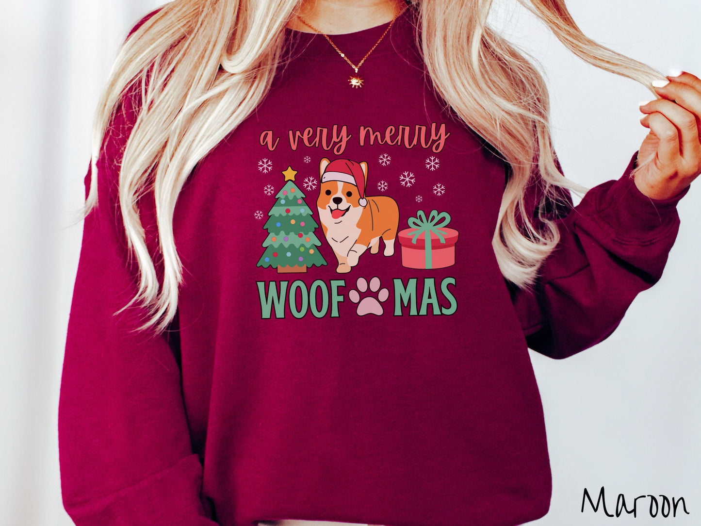A vintage maroon colored Gildan sweatshirt with the text 'a very merry Woofmas' in red and green font, featuring an orange corgi wearing a red Santa hat sitting between a Christmas tree and a present with white snowflakes hanging in the air.