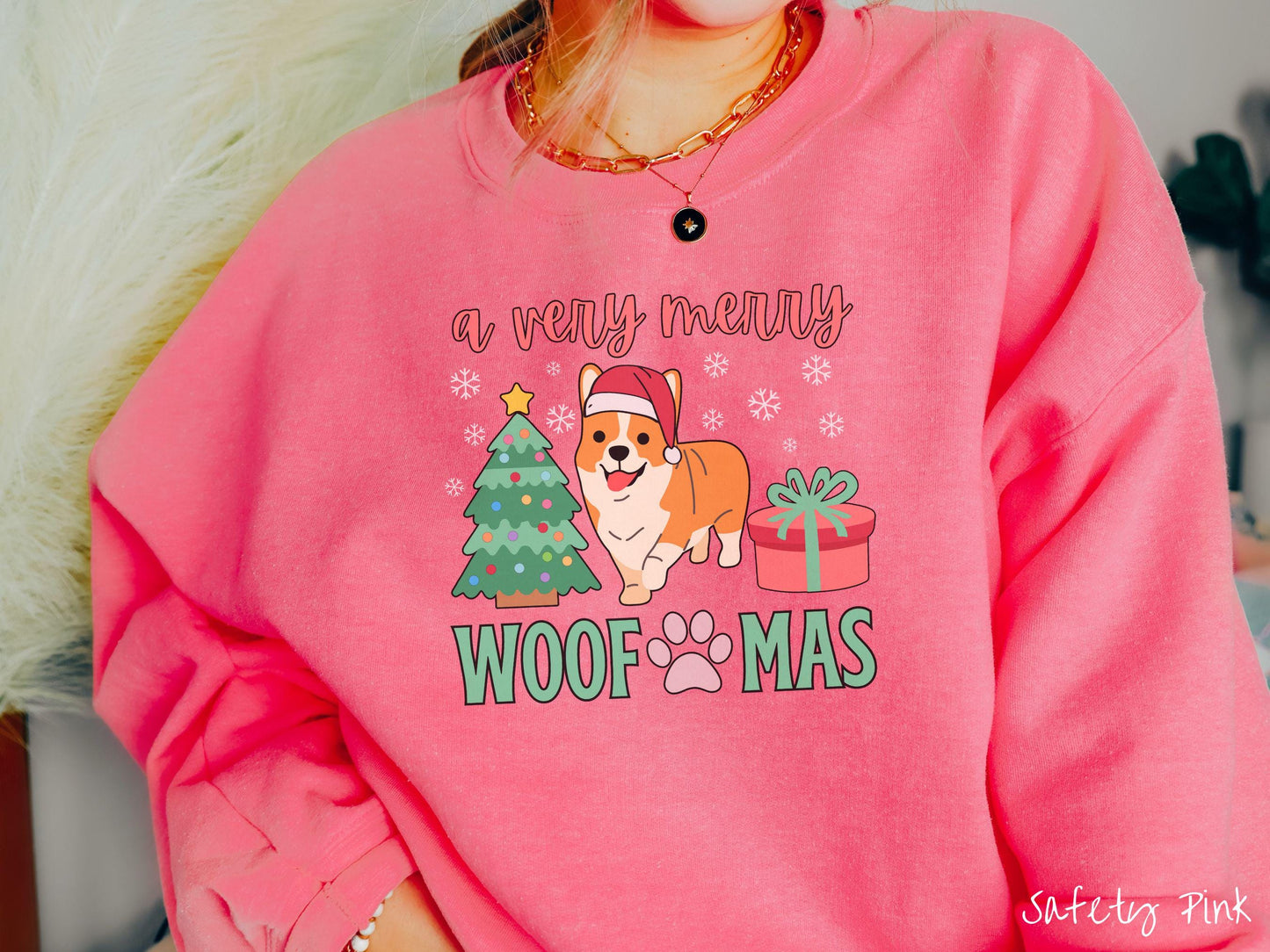 A vintage safety pink colored Gildan sweatshirt with the text 'a very merry Woofmas' in red and green font, featuring an orange corgi wearing a red Santa hat sitting between a Christmas tree and a present with white snowflakes hanging in the air.