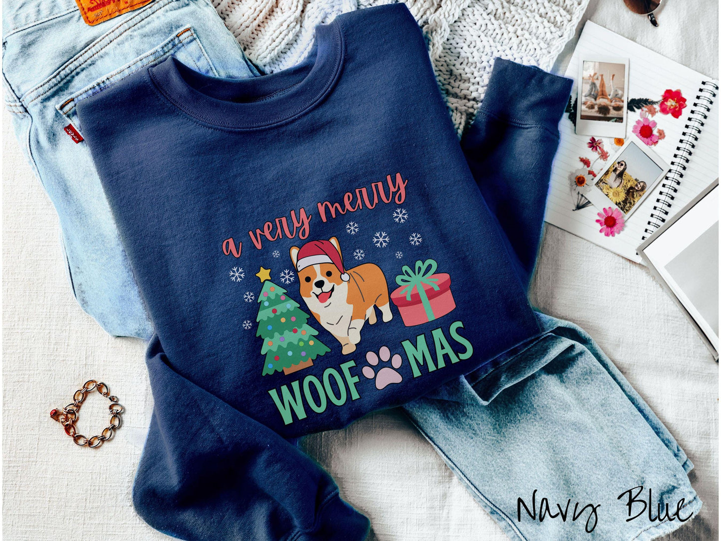 A vintage navy blue colored Gildan sweatshirt with the text 'a very merry Woofmas' in red and green font, featuring an orange corgi wearing a red Santa hat sitting between a Christmas tree and a present with white snowflakes hanging in the air.