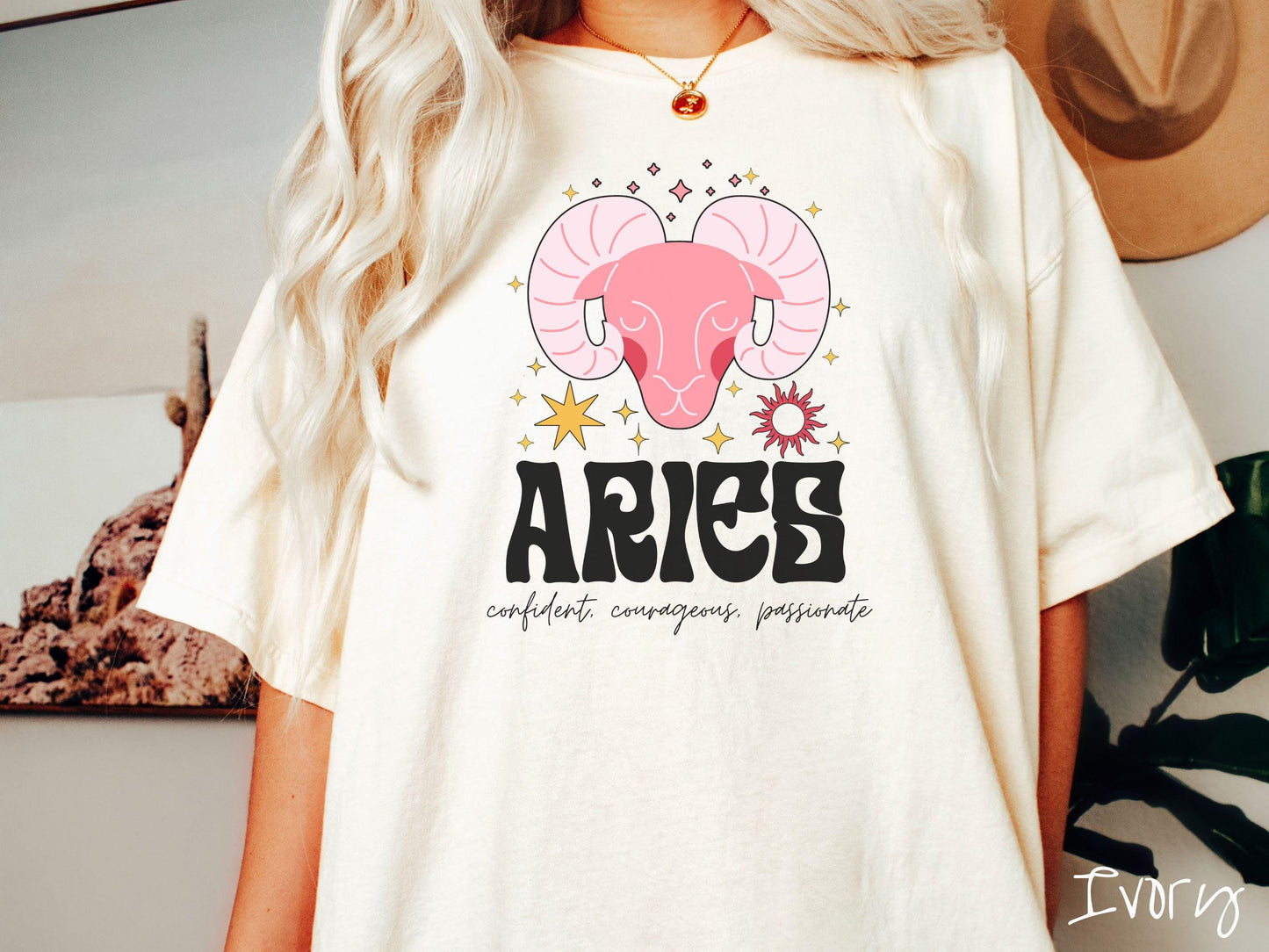 A vintage ivory colored Comfort Colors T-shirt features a large red ram head surorunded by stars and the sun with the text Aries in large bold font and below that ar the words confident, courageous passionate in cursive font.