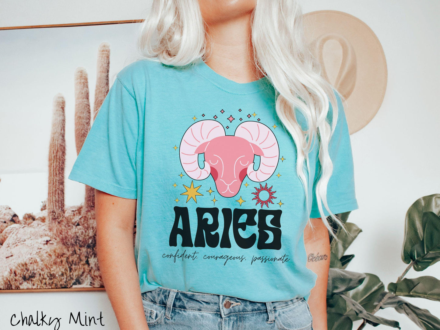 A vintage chalky mint colored Comfort Colors T-shirt features a large red ram head surorunded by stars and the sun with the text Aries in large bold font and below that ar the words confident, courageous passionate in cursive font.