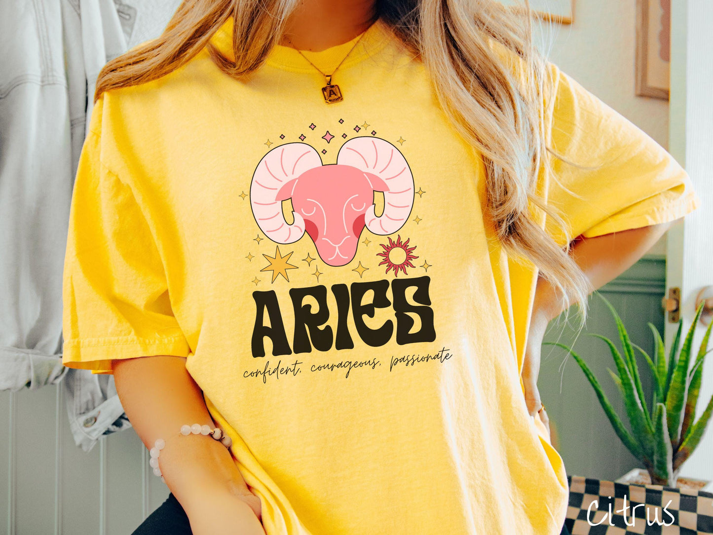 A vintage citrus colored Comfort Colors T-shirt features a large red ram head surorunded by stars and the sun with the text Aries in large bold font and below that ar the words confident, courageous passionate in cursive font.