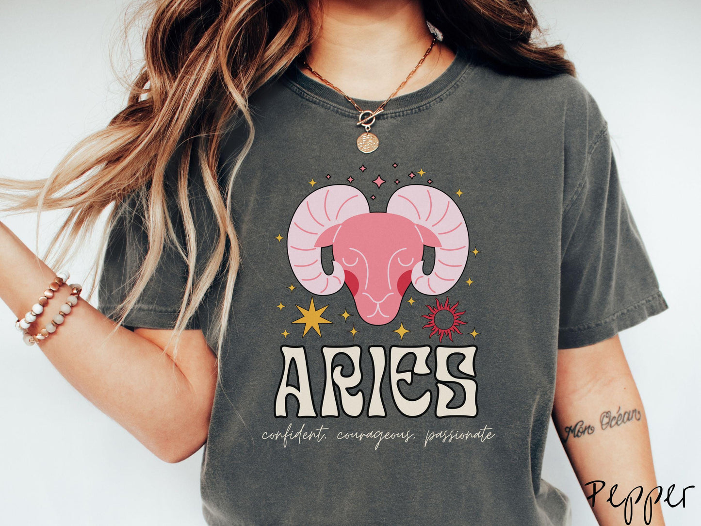 A vintage pepper colored Comfort Colors T-shirt features a large red ram head surorunded by stars and the sun with the text Aries in large bold font and below that ar the words confident, courageous passionate in cursive font.