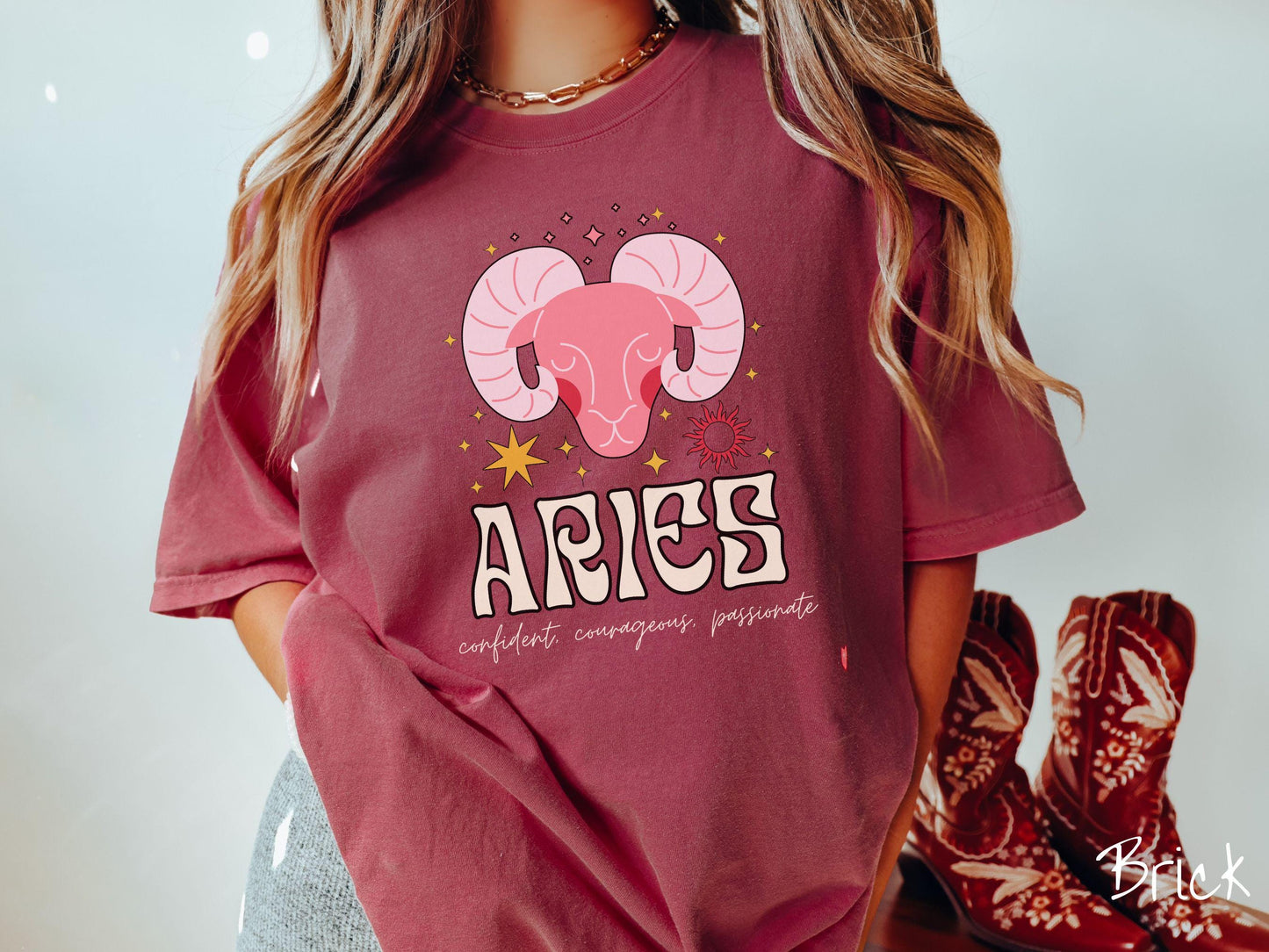 A vintage brick colored Comfort Colors T-shirt features a large red ram head surorunded by stars and the sun with the text Aries in large bold font and below that ar the words confident, courageous passionate in cursive font.