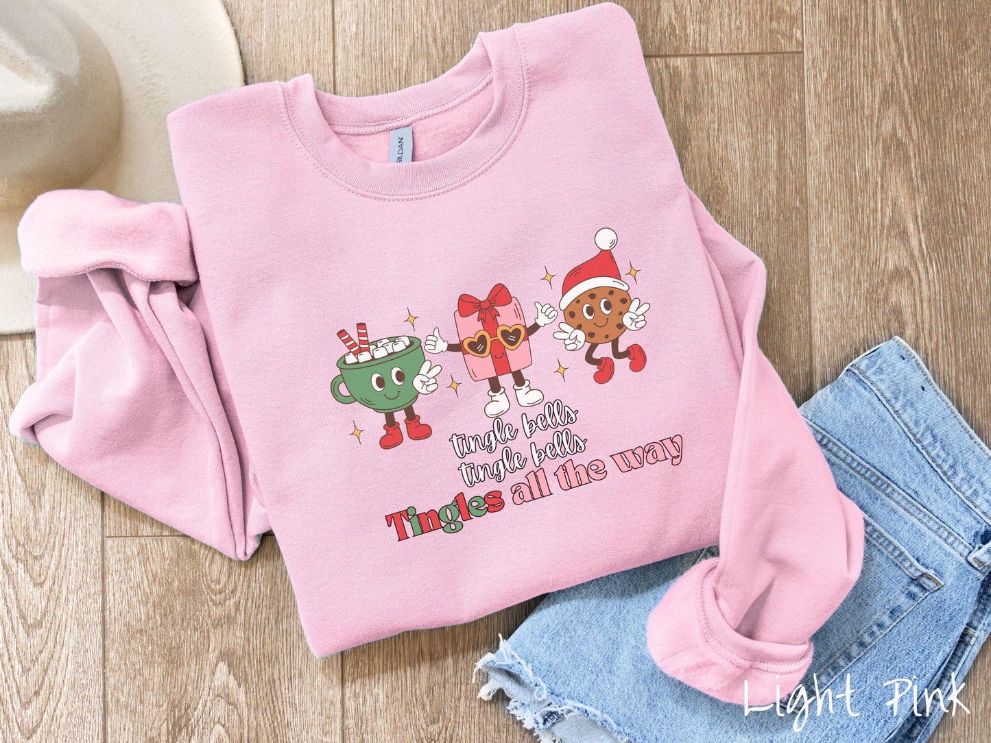 A cute, vintage light pink colored Gildan sweatshirt with text tingle bells, tingle bells, Tingles all the way. Above are a green cup of hot cocoa, a red and pink present wearing heart-shaped glasses, and a chocolate chip cookie wearing a Santa hat.
