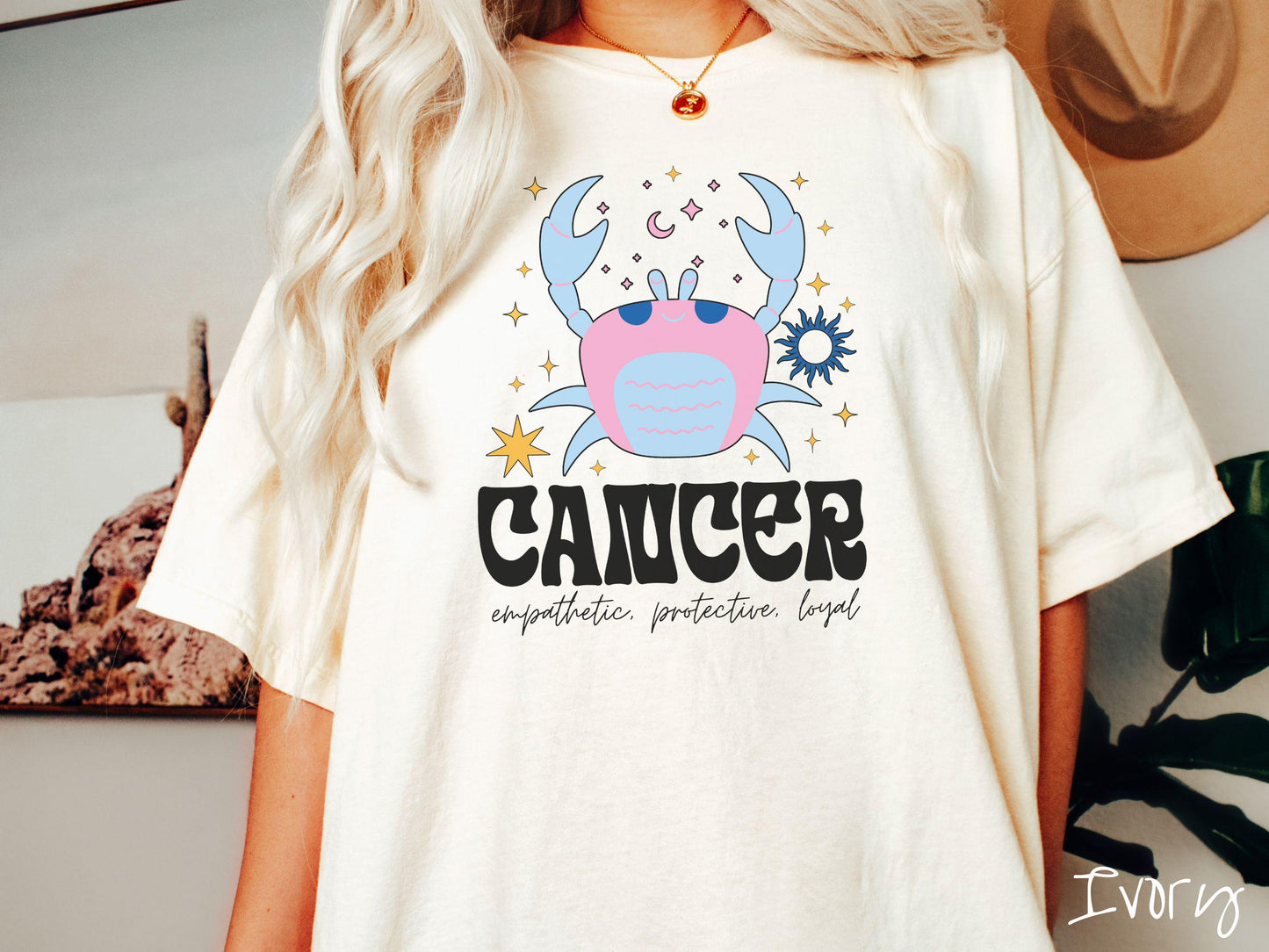 A vintage ivory colored Comfort Colors T-shirt features a large blue and pink crab surorunded by stars and the sun with the text Cancer in large bold font and below that ar the words empathetic, protective, and loyal in cursive font.
