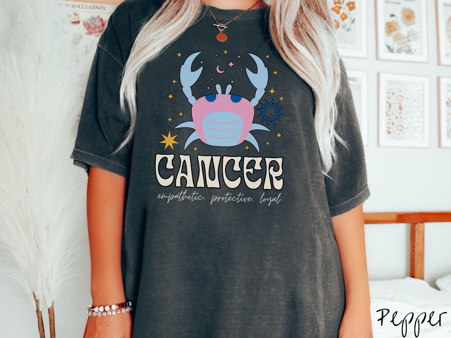 A vintage pepper colored Comfort Colors T-shirt features a large blue and pink crab surorunded by stars and the sun with the text Cancer in large bold font and below that ar the words empathetic, protective, and loyal in cursive font.