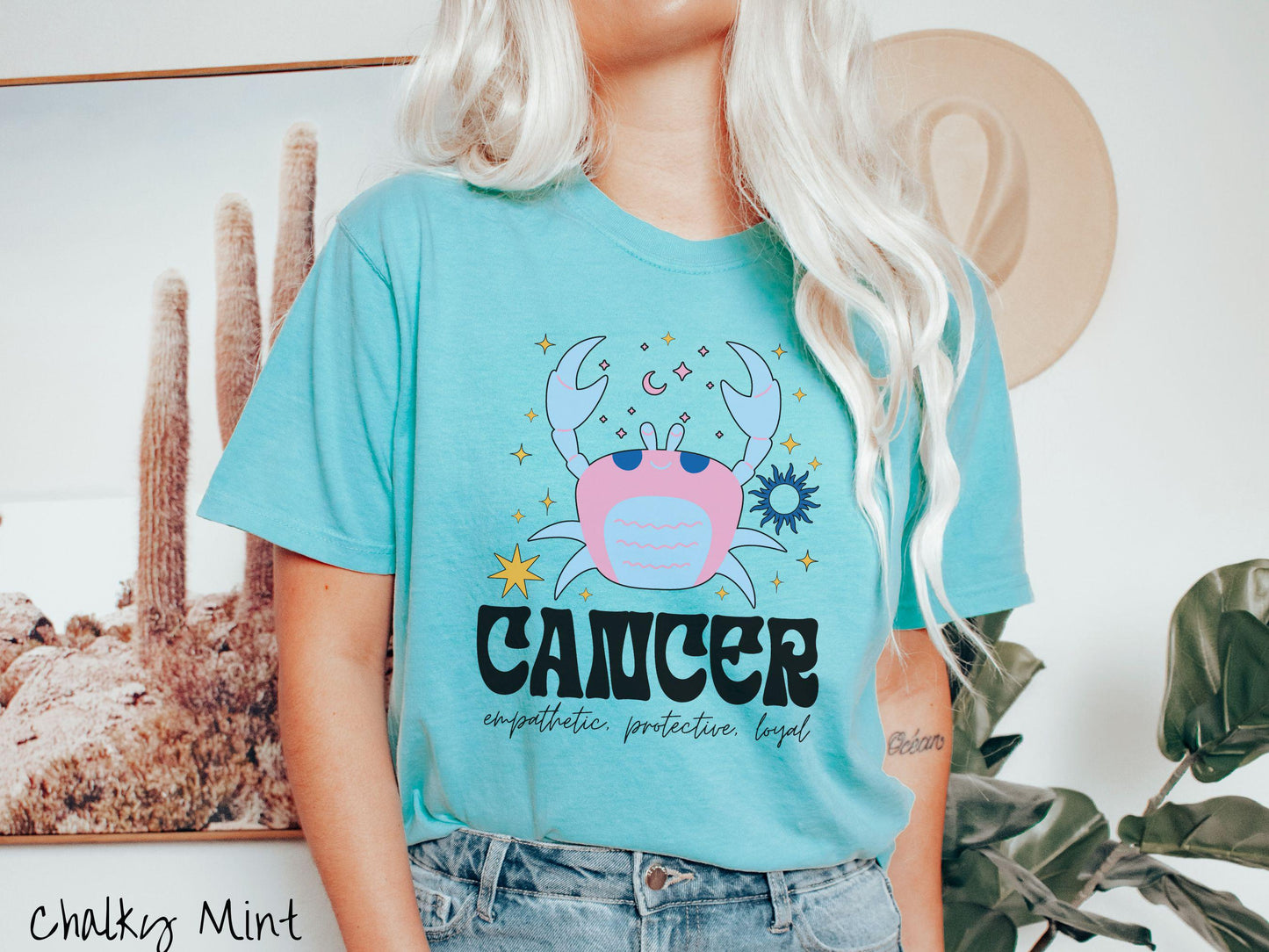 A vintage chalky mint colored Comfort Colors T-shirt features a large blue and pink crab surorunded by stars and the sun with the text Cancer in large bold font and below that ar the words empathetic, protective, and loyal in cursive font.