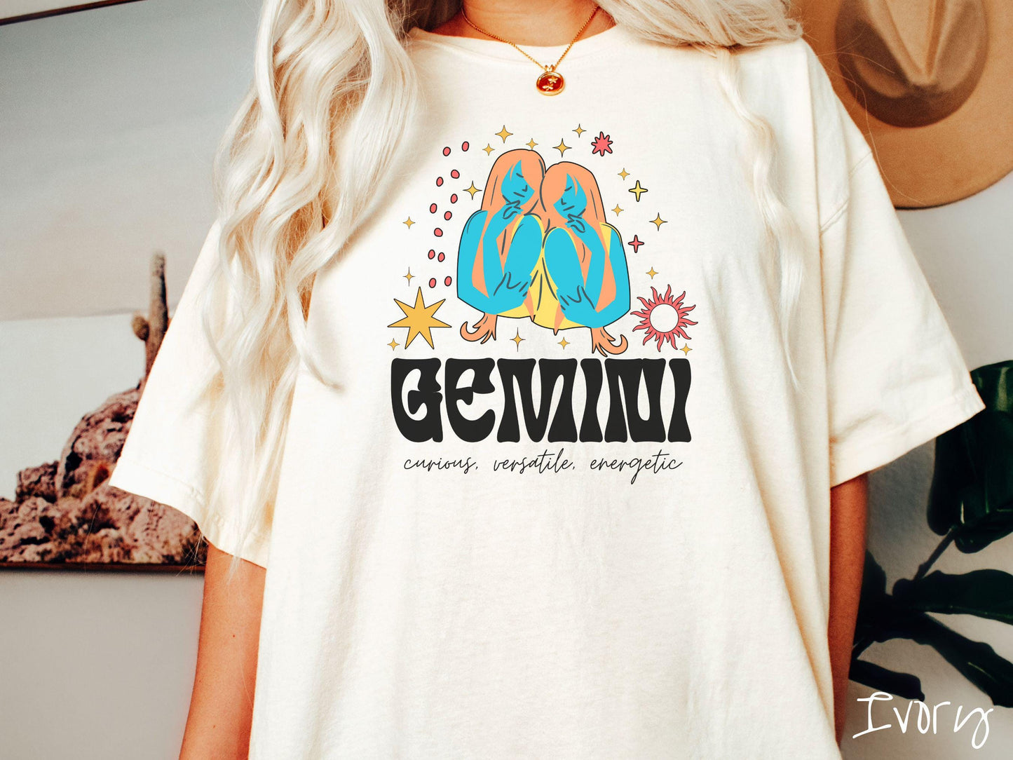 A vintage ivory colored Comfort Colors T-shirt features the twins Castor and Pollux in blue and orange colors surrounded by stars and the sun with text Gemini in large bold font and below are the words curious, versatile, energetic in cursive font.